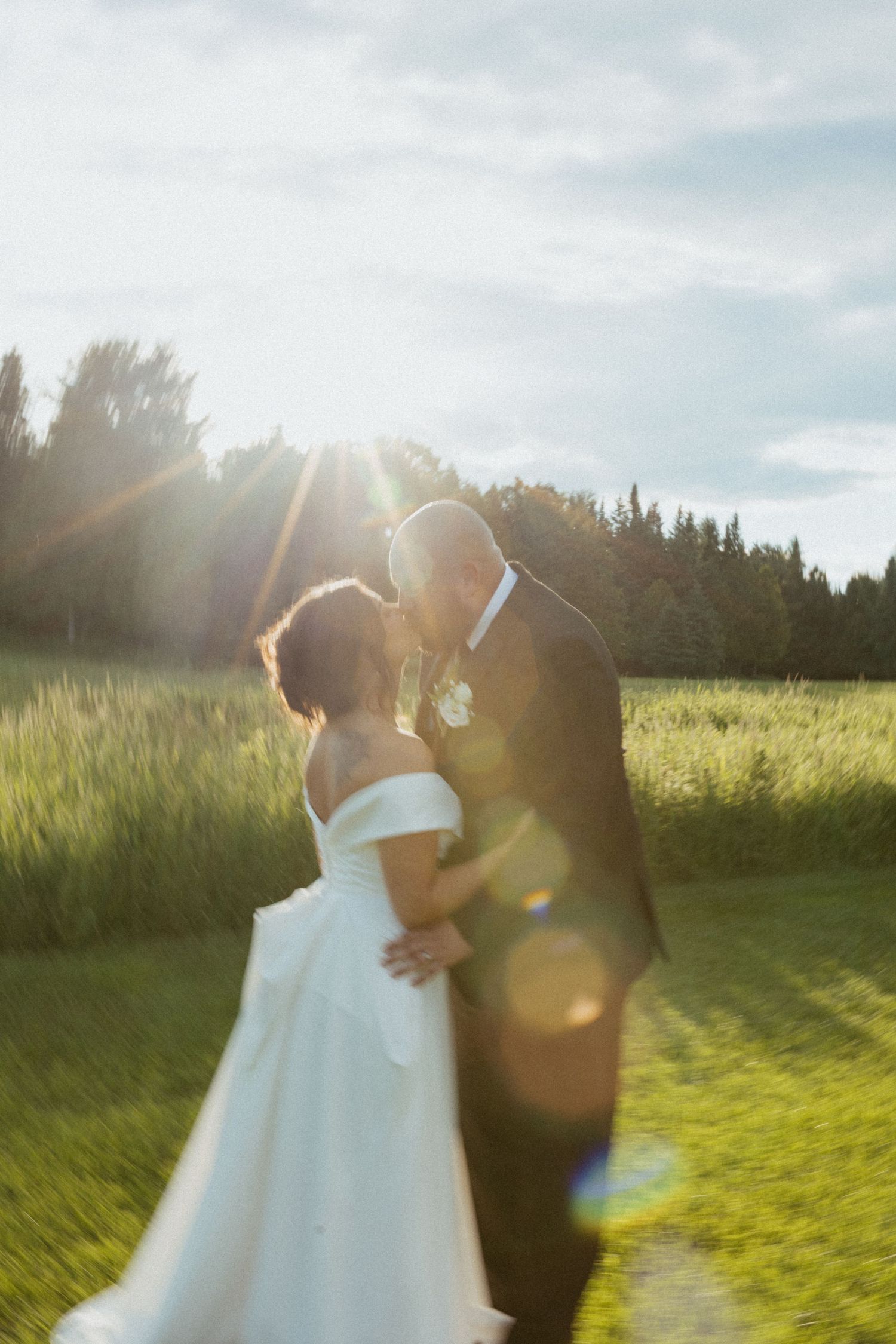 Bride and groom sharing a romantic kiss at golden hour in a lush green field with sun flare.