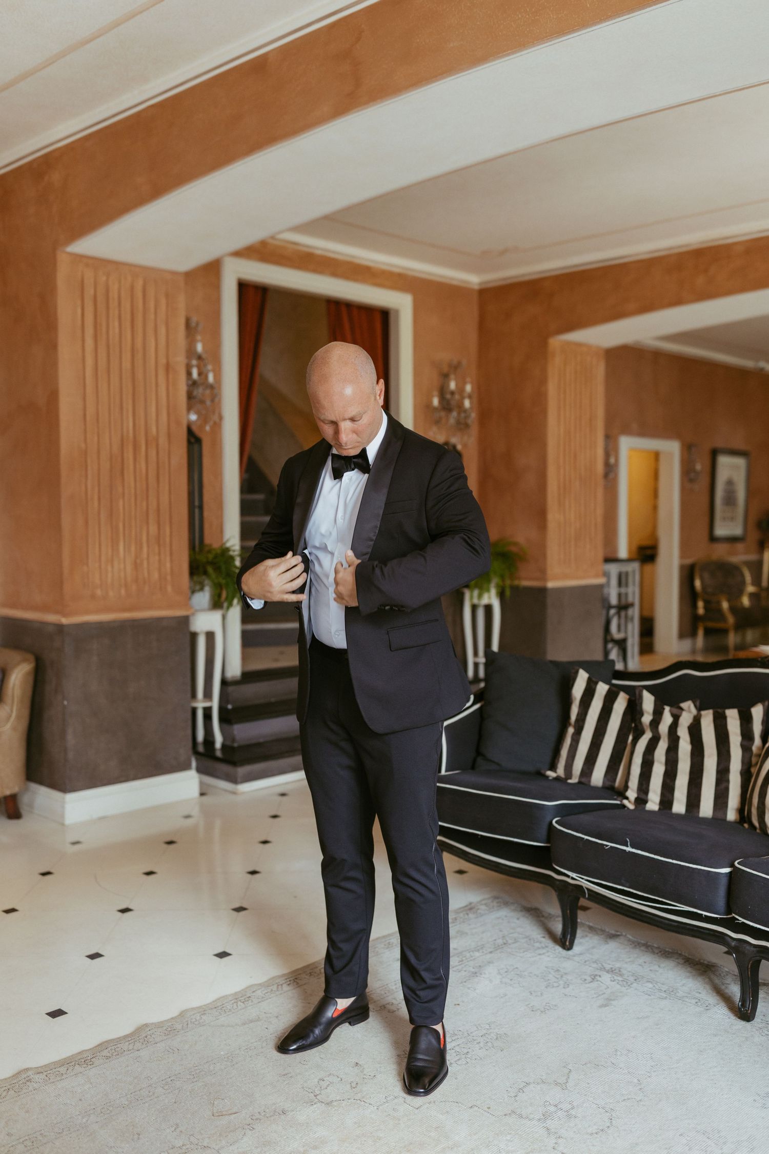 Bald groom in black tuxedo and bow tie adjusting his jacket in an elegant hotel lobby.