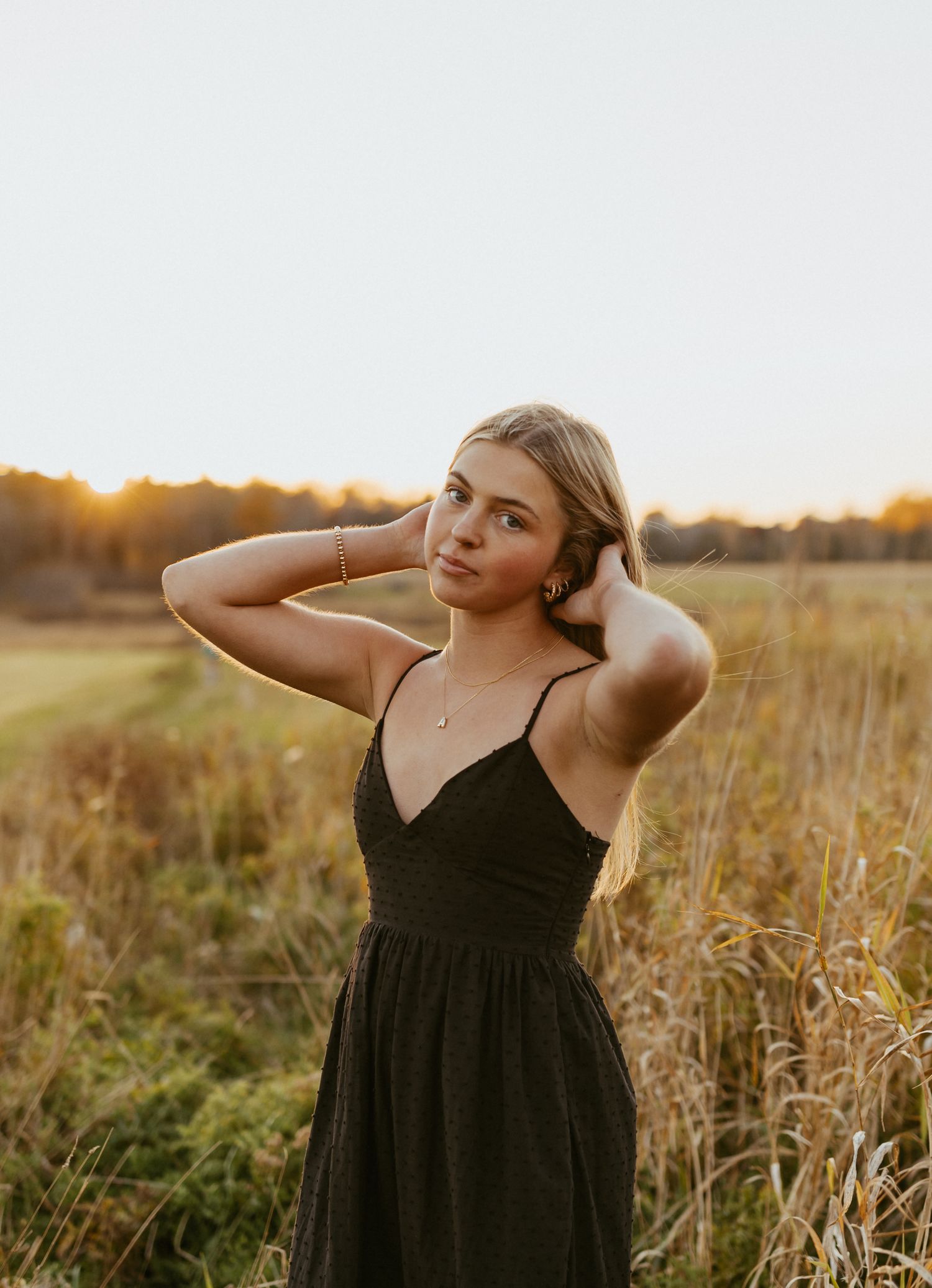 Young blonde woman in black sundress poses gracefully in golden field at sunset.