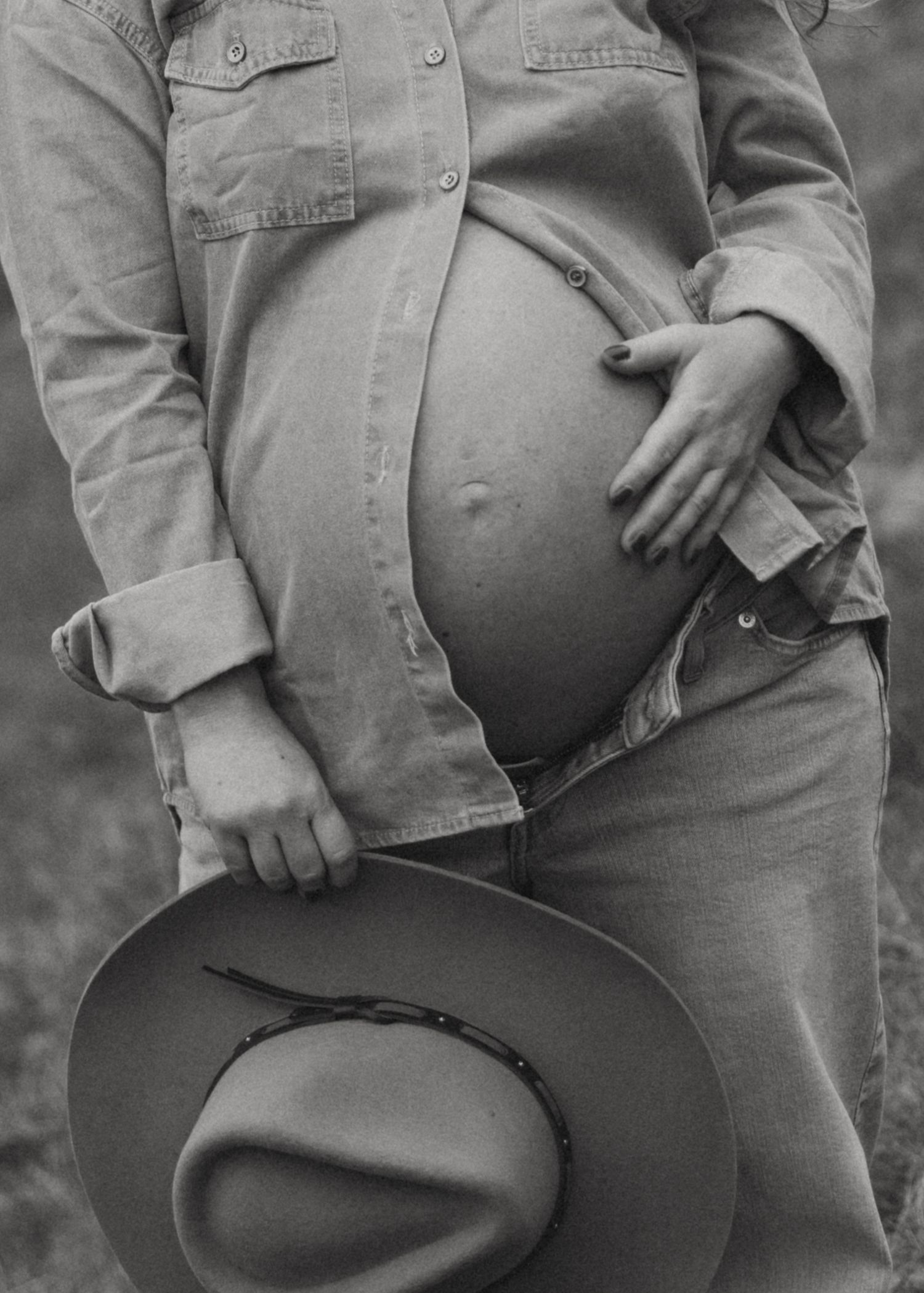 Black and white photo of pregnant woman in denim shirt and jeans holding cowboy hat, cradling baby bump.