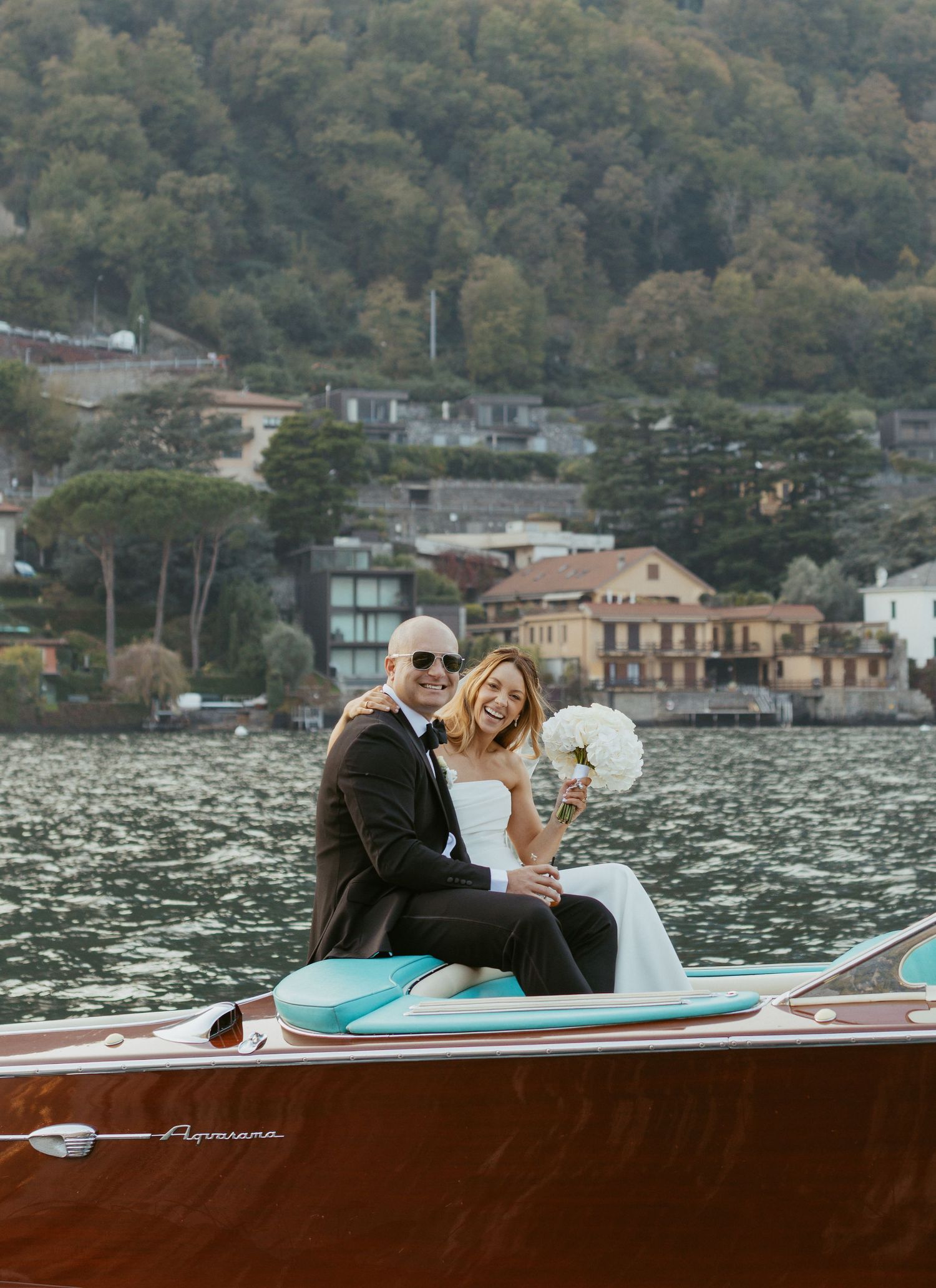 Newlyweds pose on a classic wooden Aquarama speedboat on Lake Como with a scenic Italian village backdrop.
