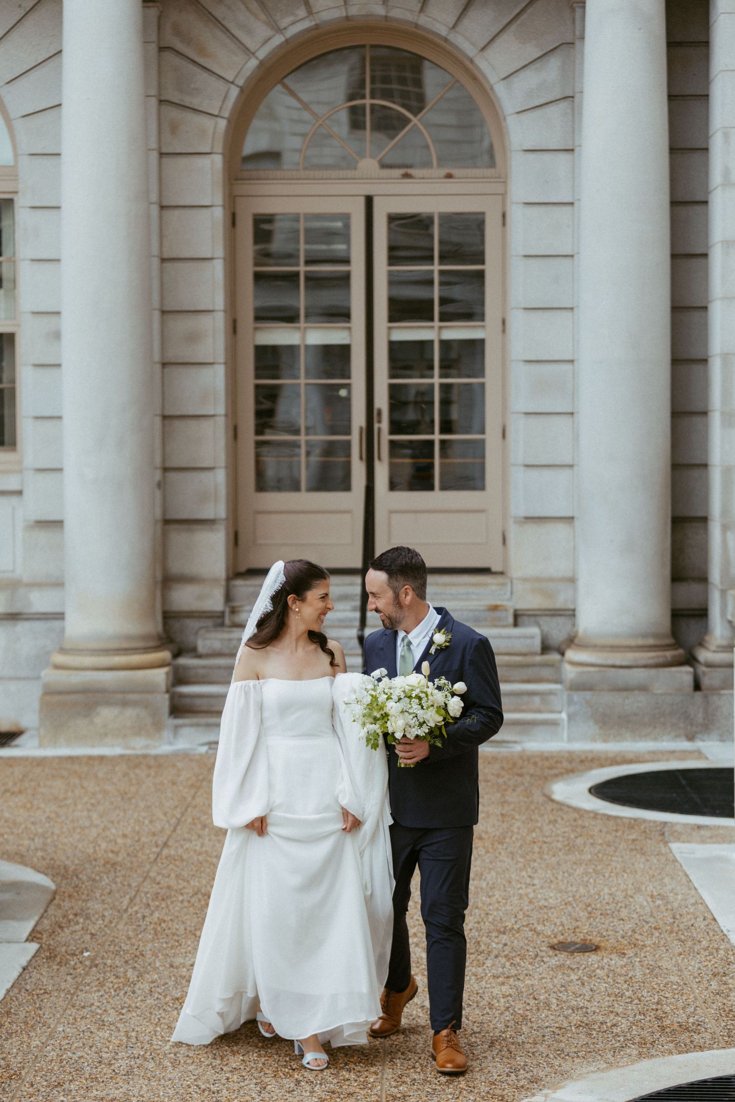 Bride and groom walk together laughing outside a neoclassical building with tall white columns.