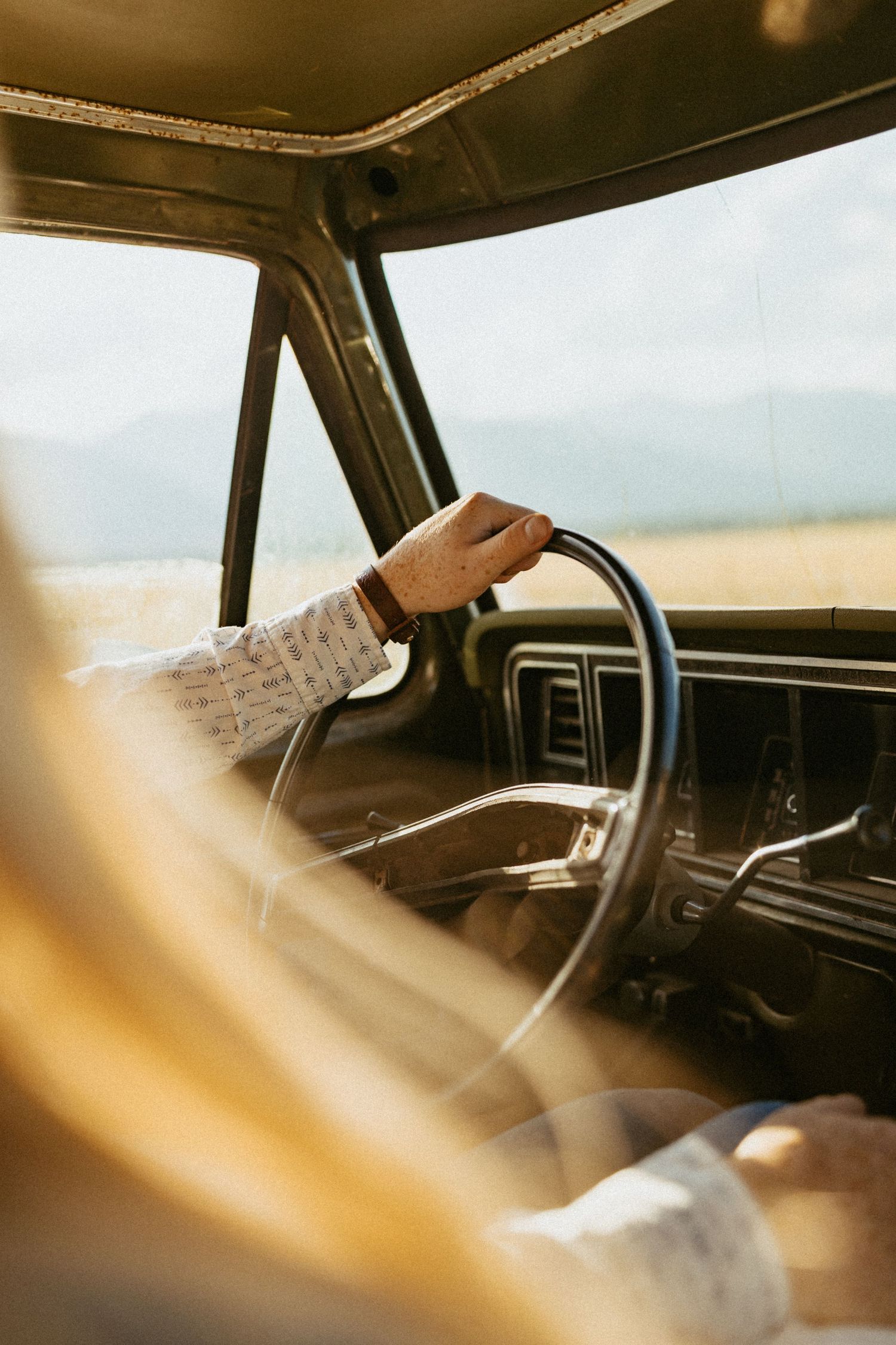 Person gripping vintage truck steering wheel wearing patterned shirt with golden sunlit mountains in background.