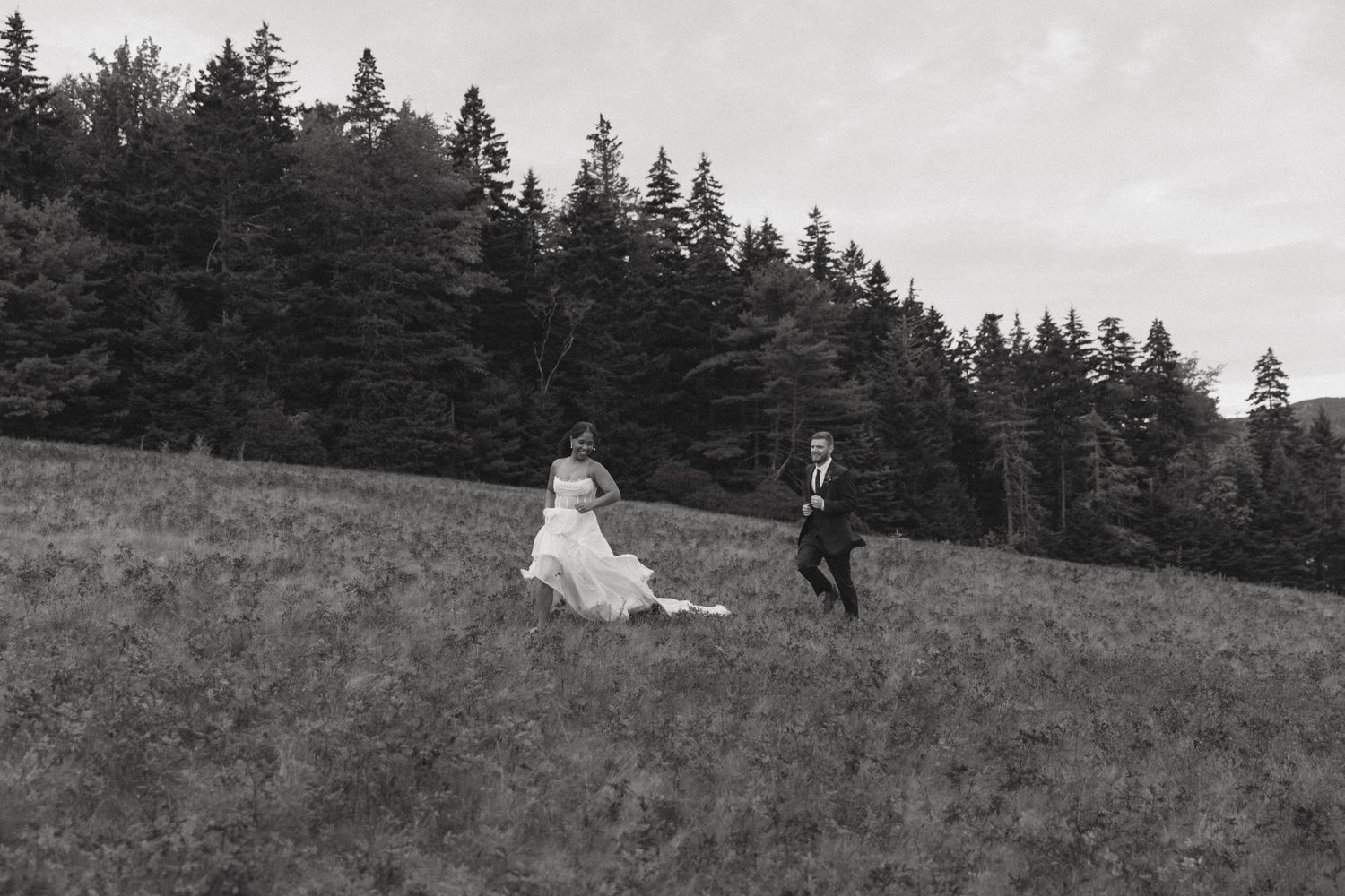 Black and white wedding photo of couple running across a hillside meadow with evergreen trees.