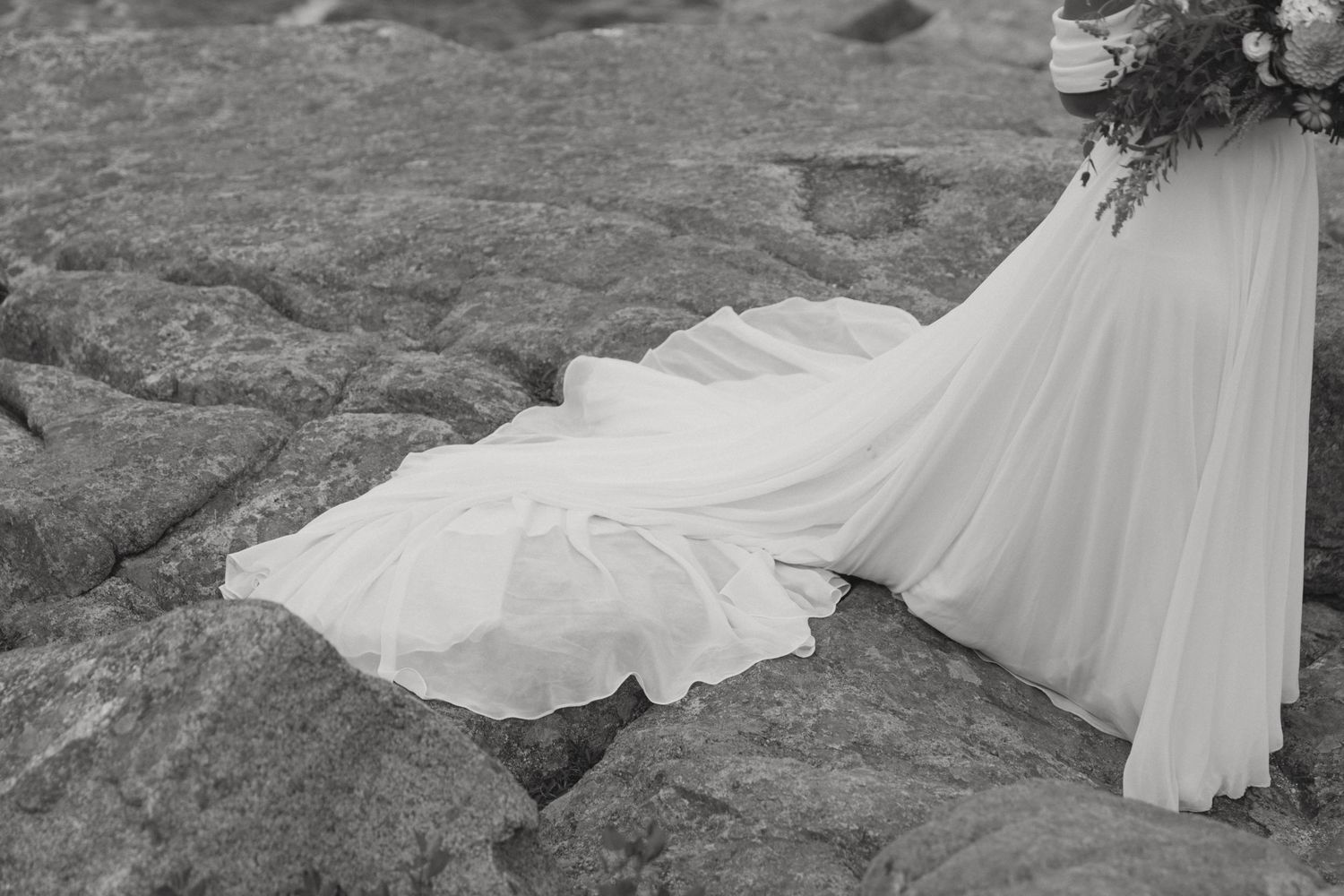 Black and white close-up of a flowing bridal gown train on rocky coastal terrain.