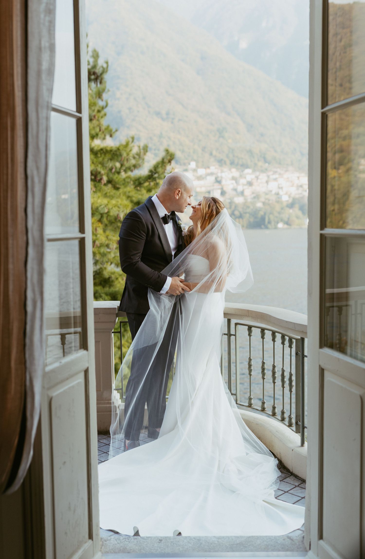 Bride and groom kissing on a scenic balcony overlooking Lake Como mountains, framed by elegant French doors.