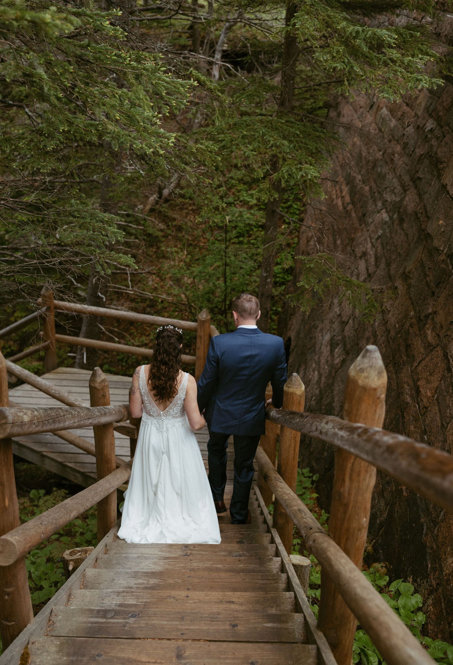 Bride and groom walking hand-in-hand down rustic wooden stairs surrounded by lush forest on their wedding day.
