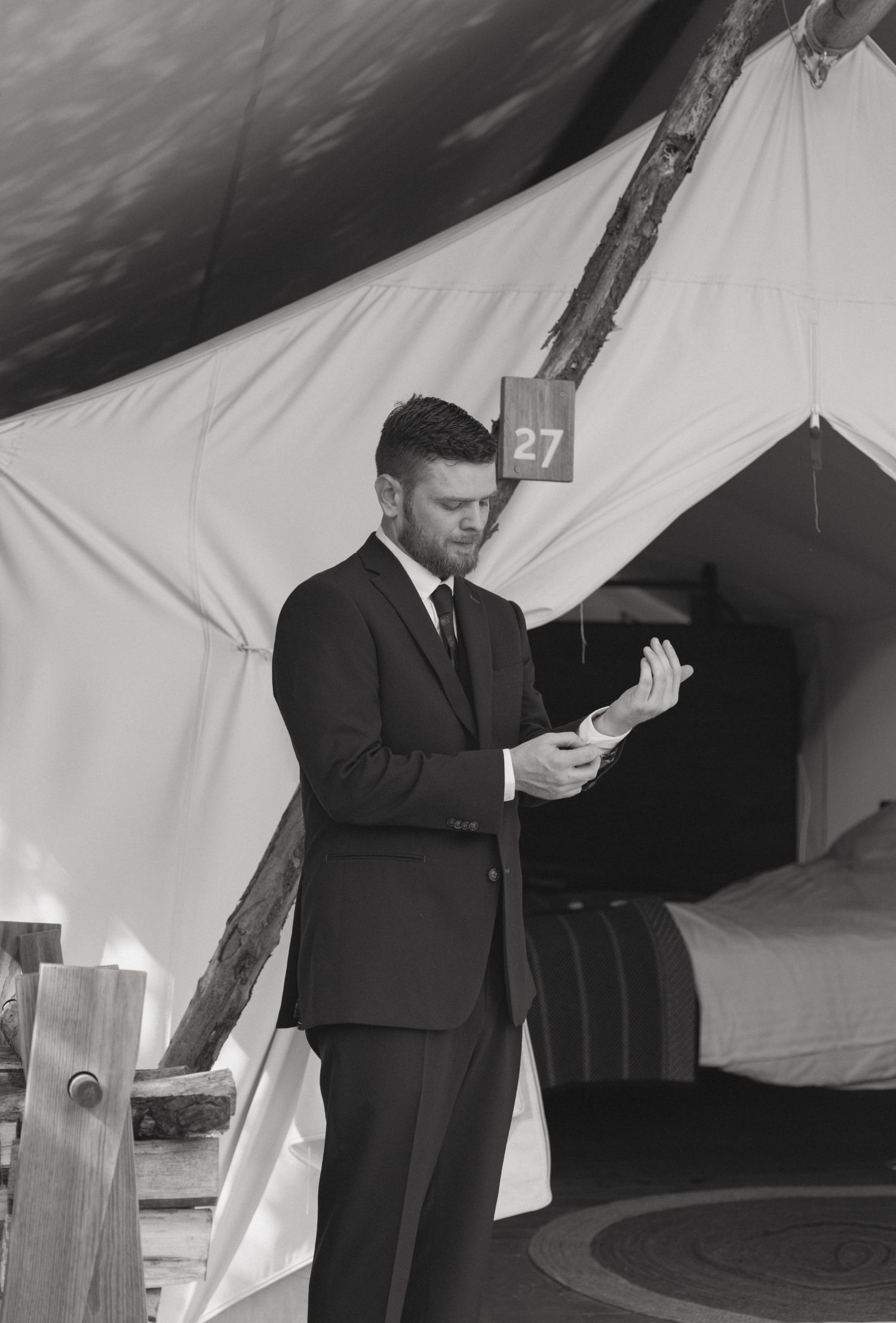 Black and white photo of groom in suit reading vows outside a canvas glamping tent numbered 27.