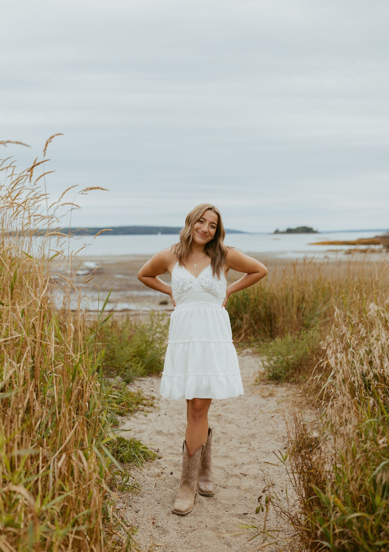 Young woman in white dress and cowboy boots poses on sandy path between tall grasses near coastal waters.