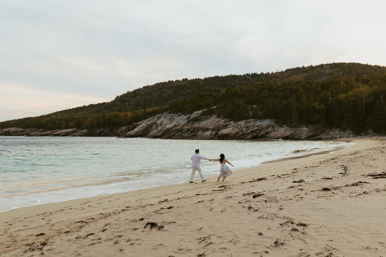 Couple holding hands walking along Acadia sandy beach with rocky cliffs and turquoise water.