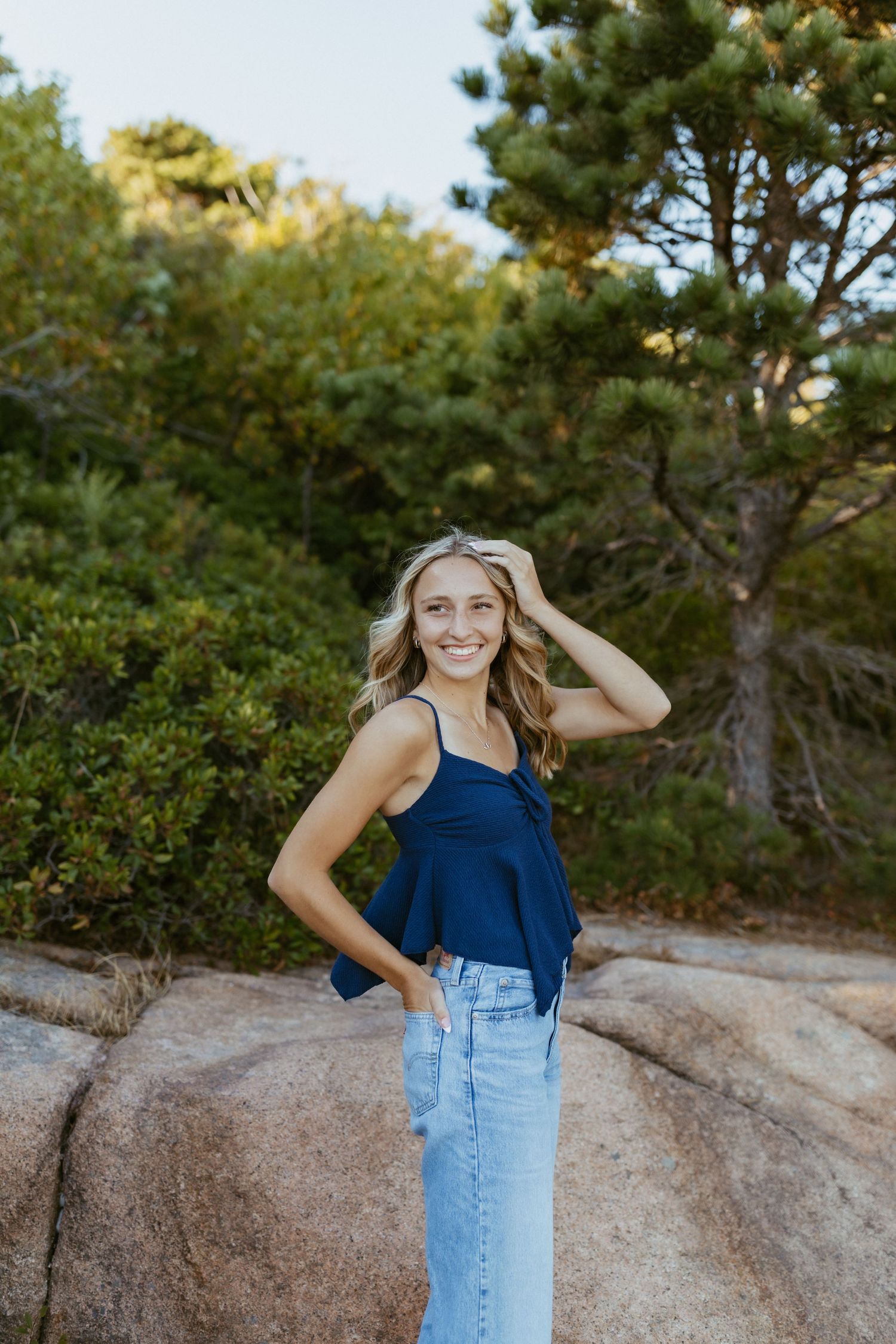Smiling blonde woman in navy top and light jeans posing on rocky terrain with lush green trees behind her.