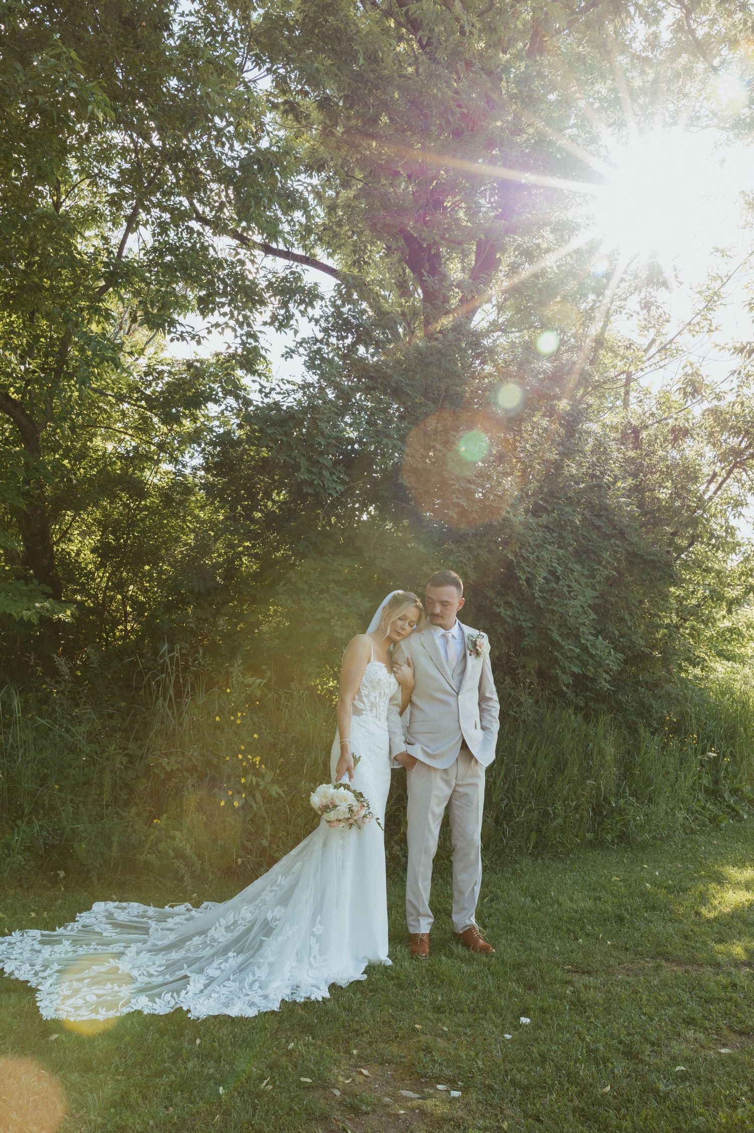 Bride and groom standing together in lush green garden with sunlight streaming through trees on their wedding day.