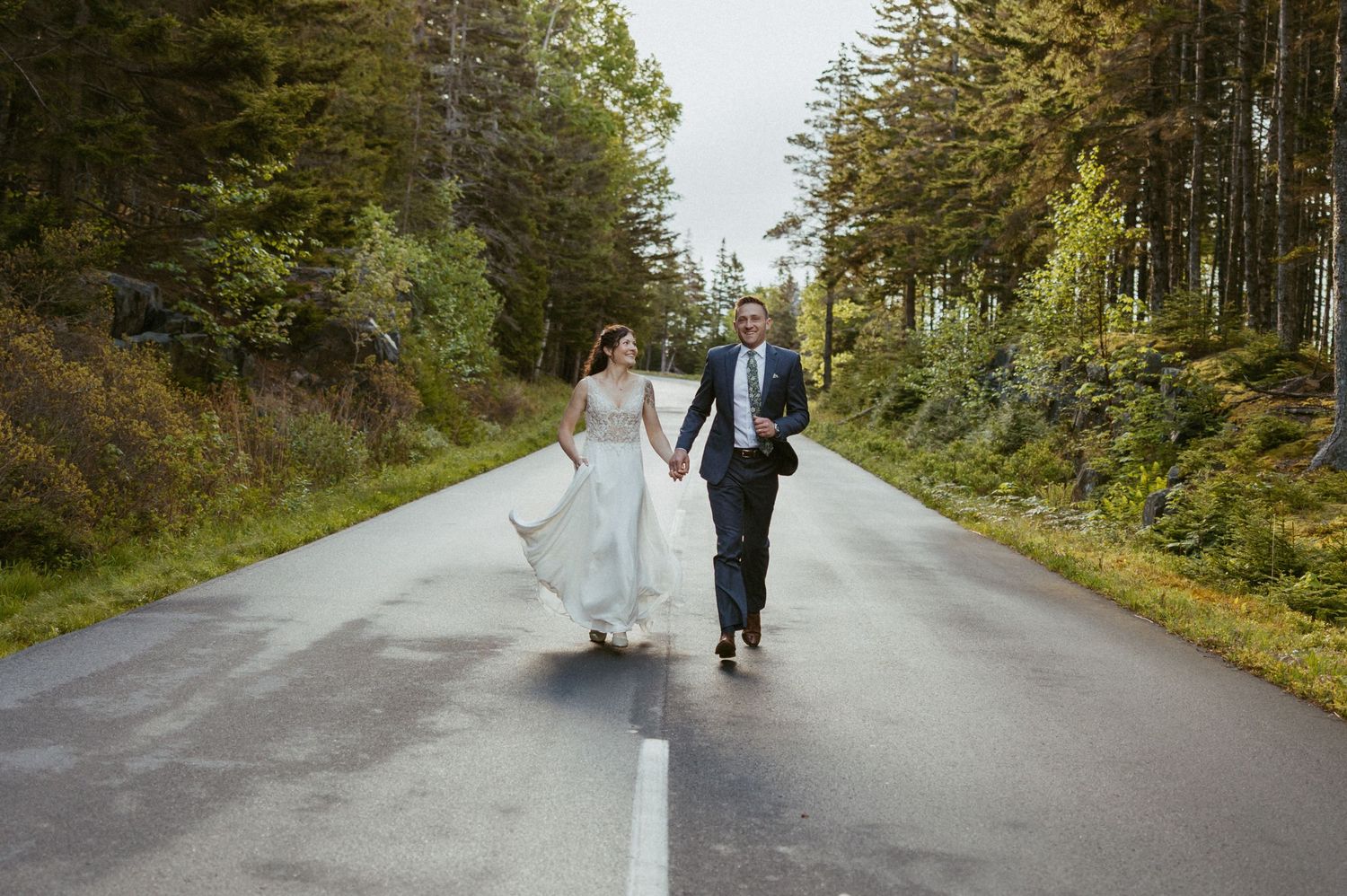 Bride and groom holding hands walking down a scenic tree-lined road in their wedding attire.