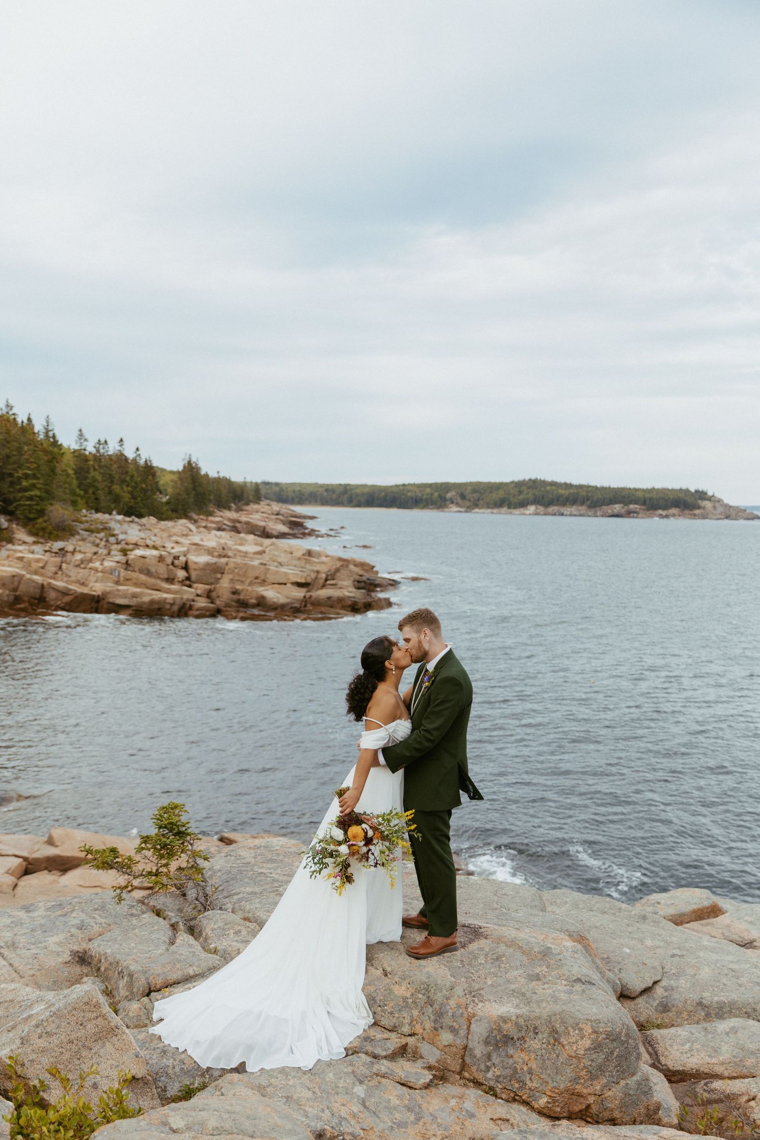 Bride and groom share a kiss on rocky coastal cliffs with ocean views at their outdoor wedding.