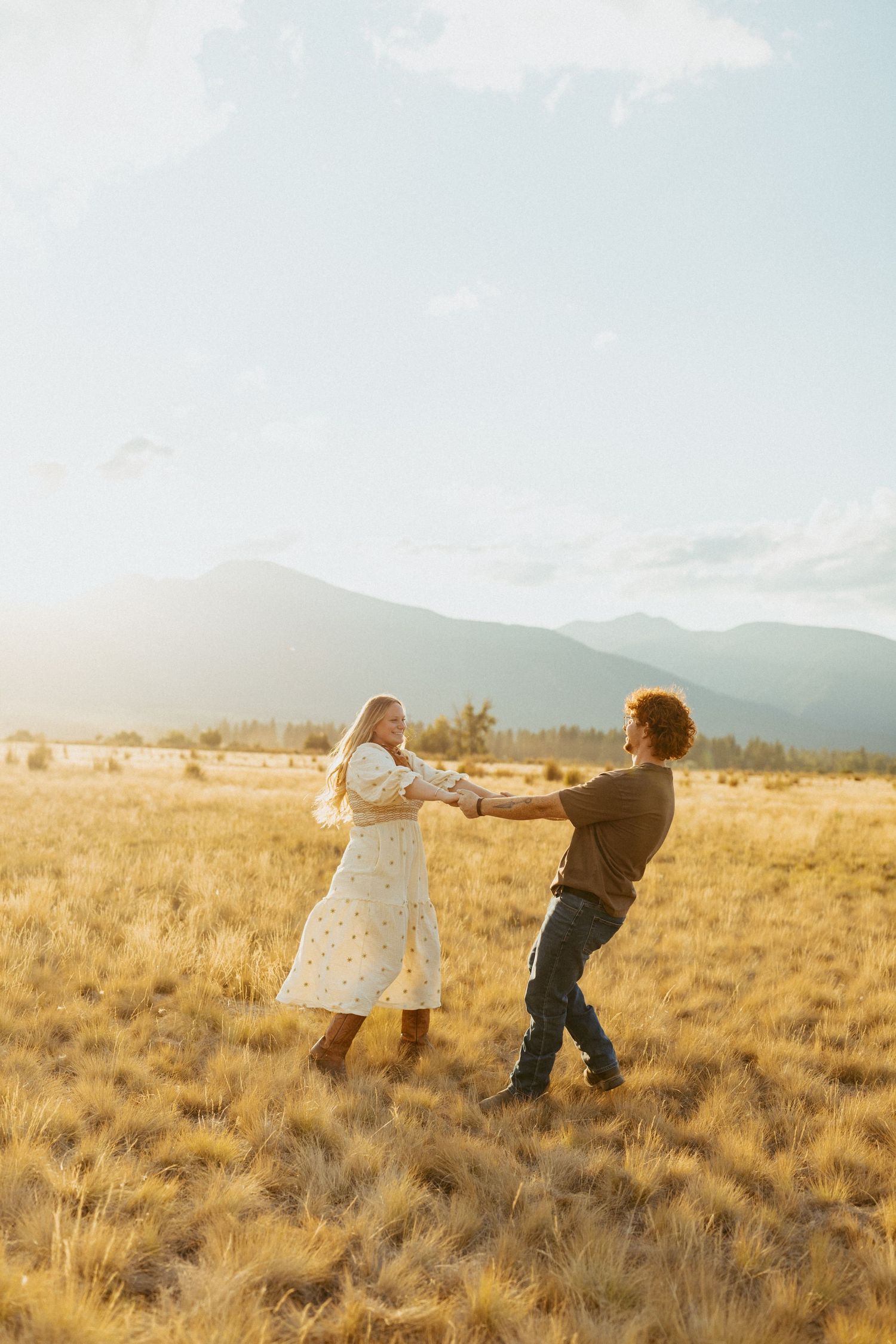 Playful couple spinning together holding hands in golden grass field with stunning Montana mountain landscape at sunset.