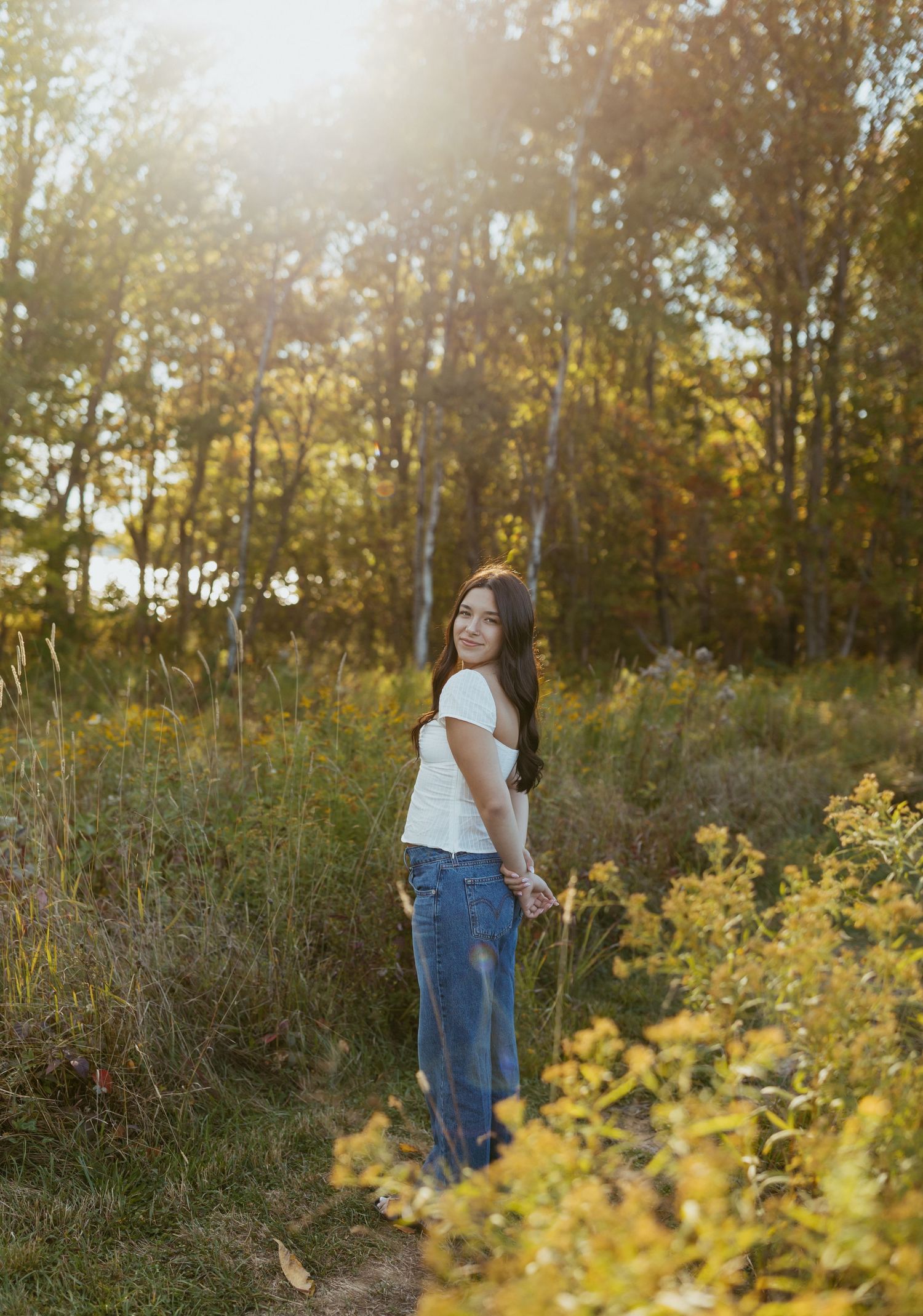 Brunette woman in white shirt and jeans standing on a sunlit forest path surrounded by fall foliage.