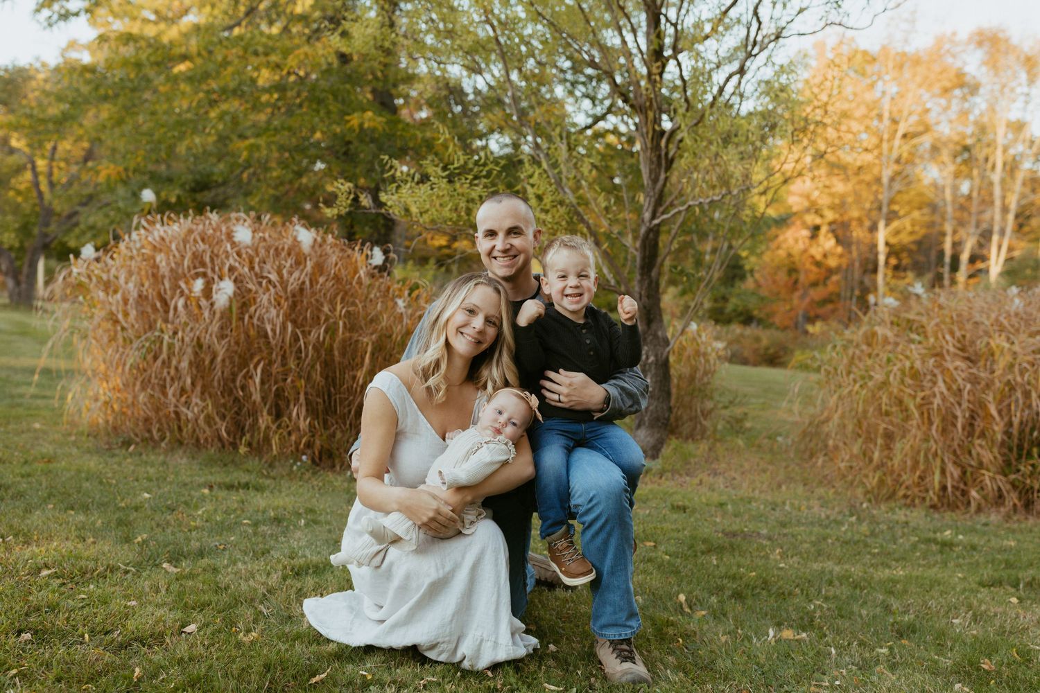Family of four posing outdoors in autumn, parents holding newborn and toddler, surrounded by fall foliage.