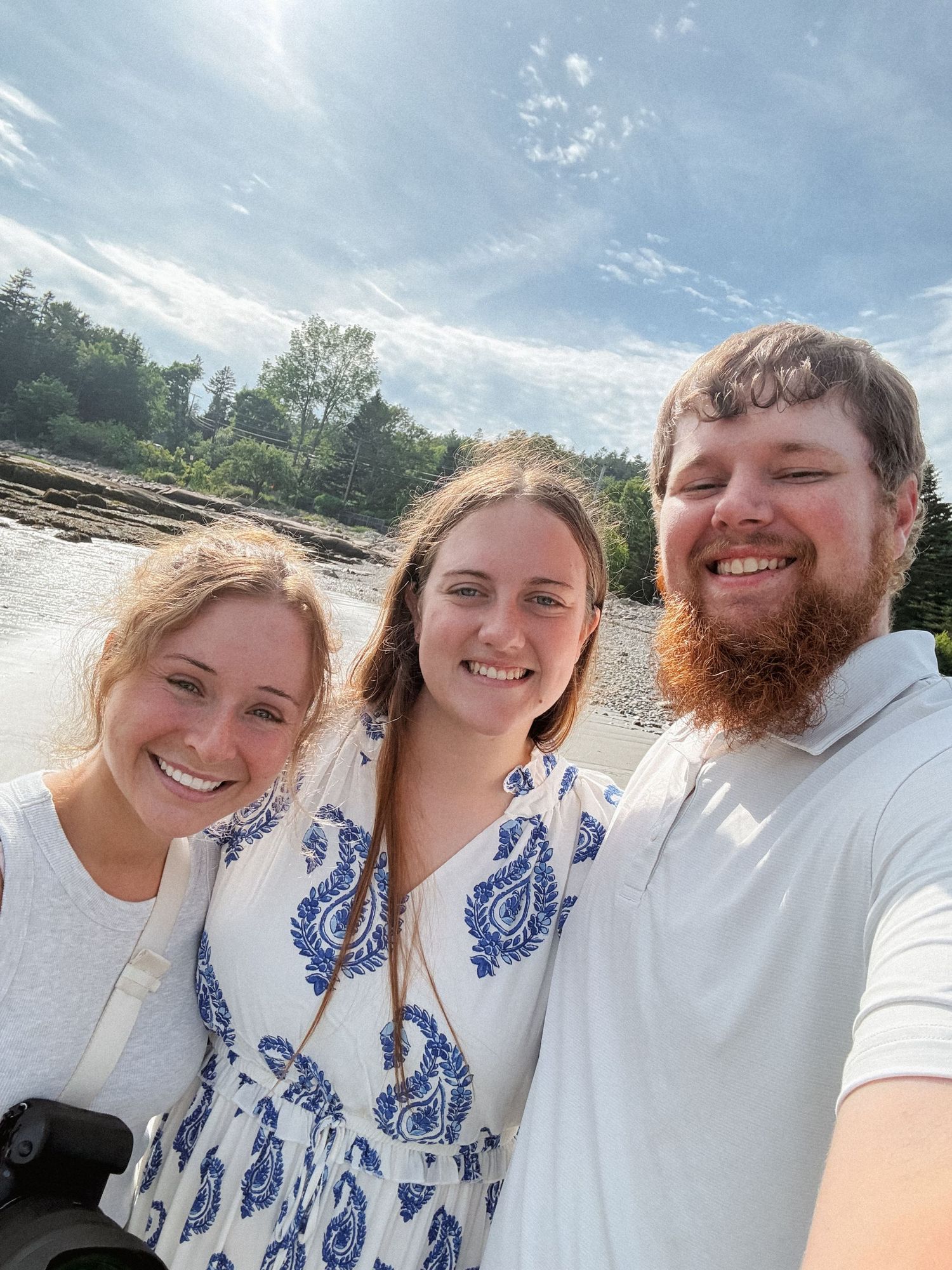 Three smiling friends pose for a selfie outdoors near a river on a sunny day with trees and blue sky behind them.