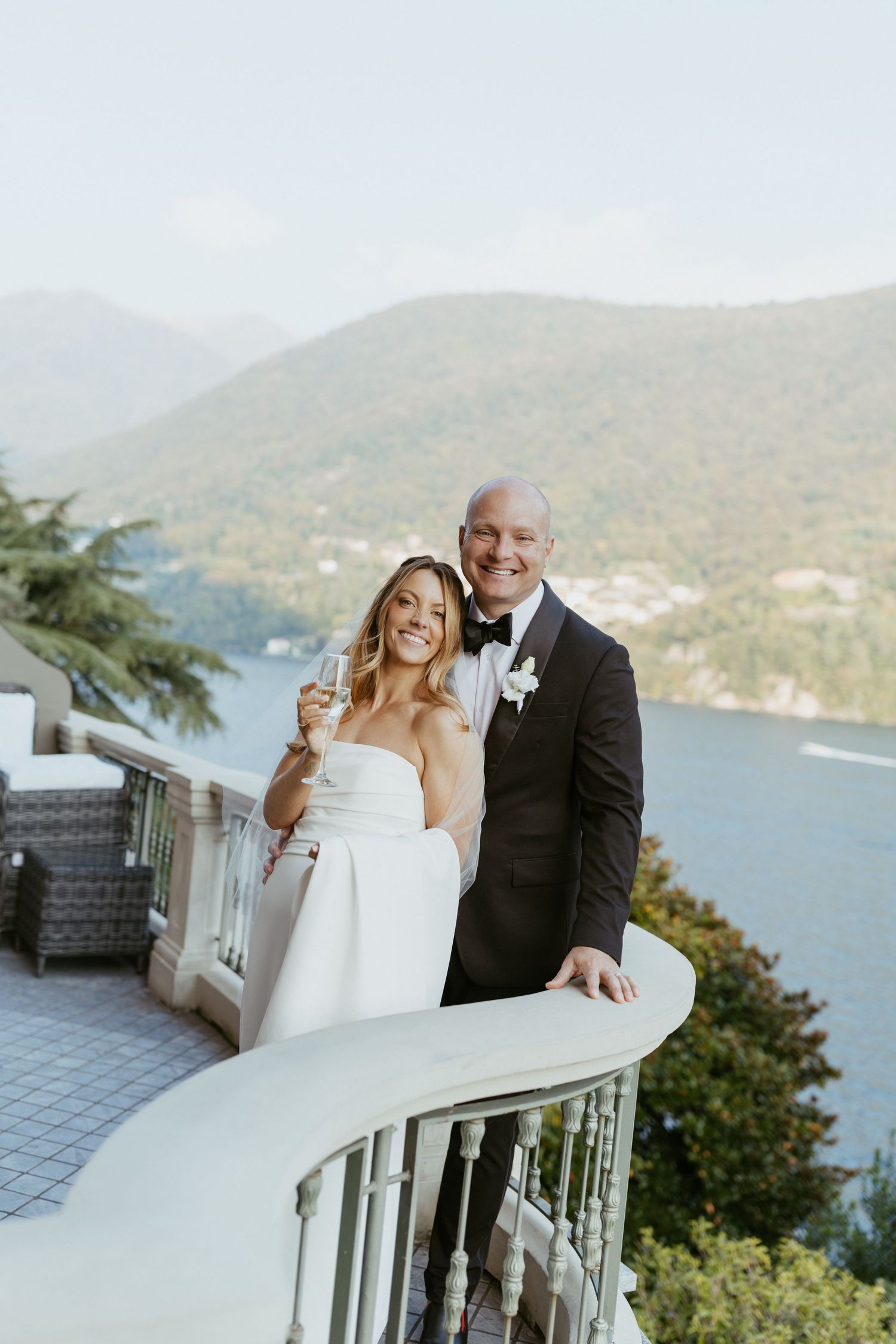 Bride and groom smiling on elegant balcony with champagne, overlooking scenic lake and mountains.