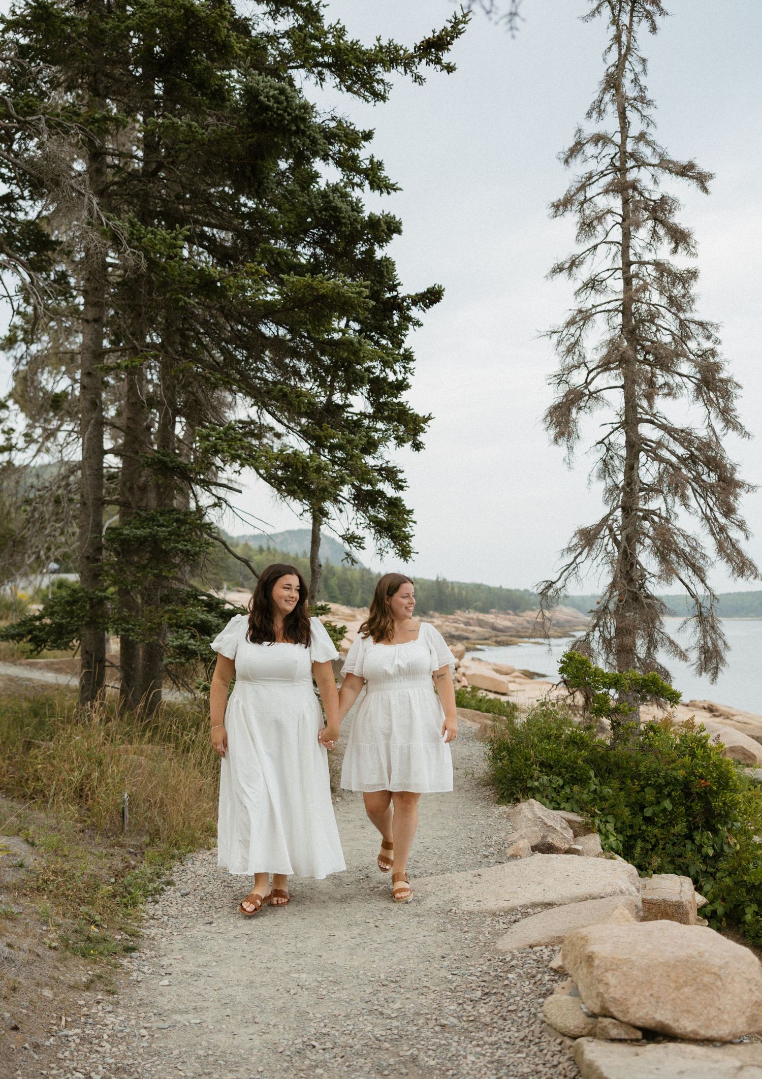 Two women in white dresses hold hands walking a scenic coastal path lined with evergreen trees and rocky shoreline.