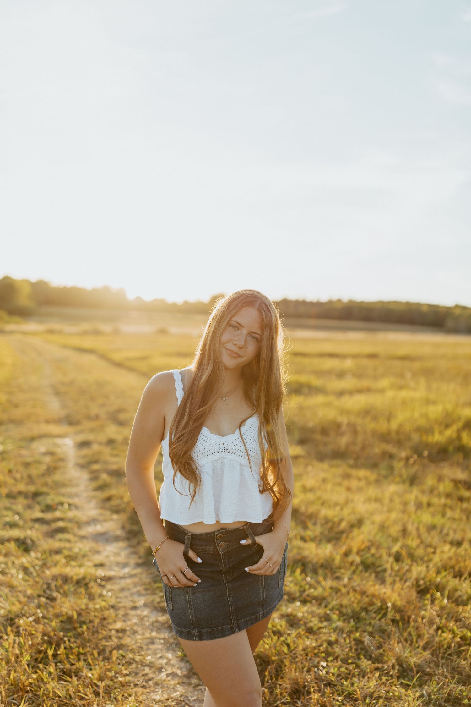High school senior in white top and denim skirt posing in golden sunlit field at sunset.