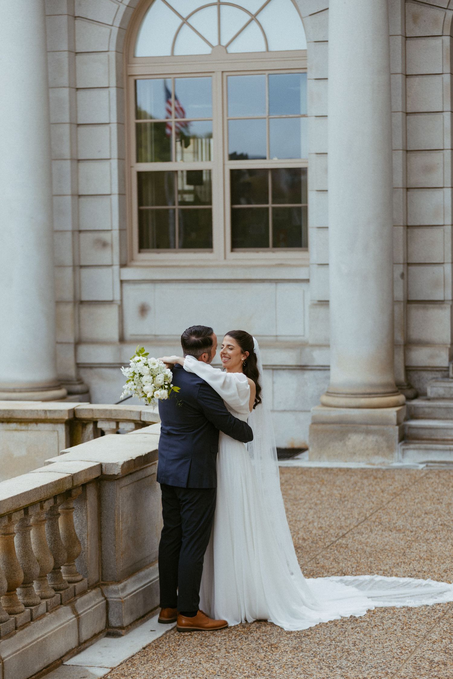 Smiling bride in long-sleeve gown embraces groom before grand white columned building exterior.
