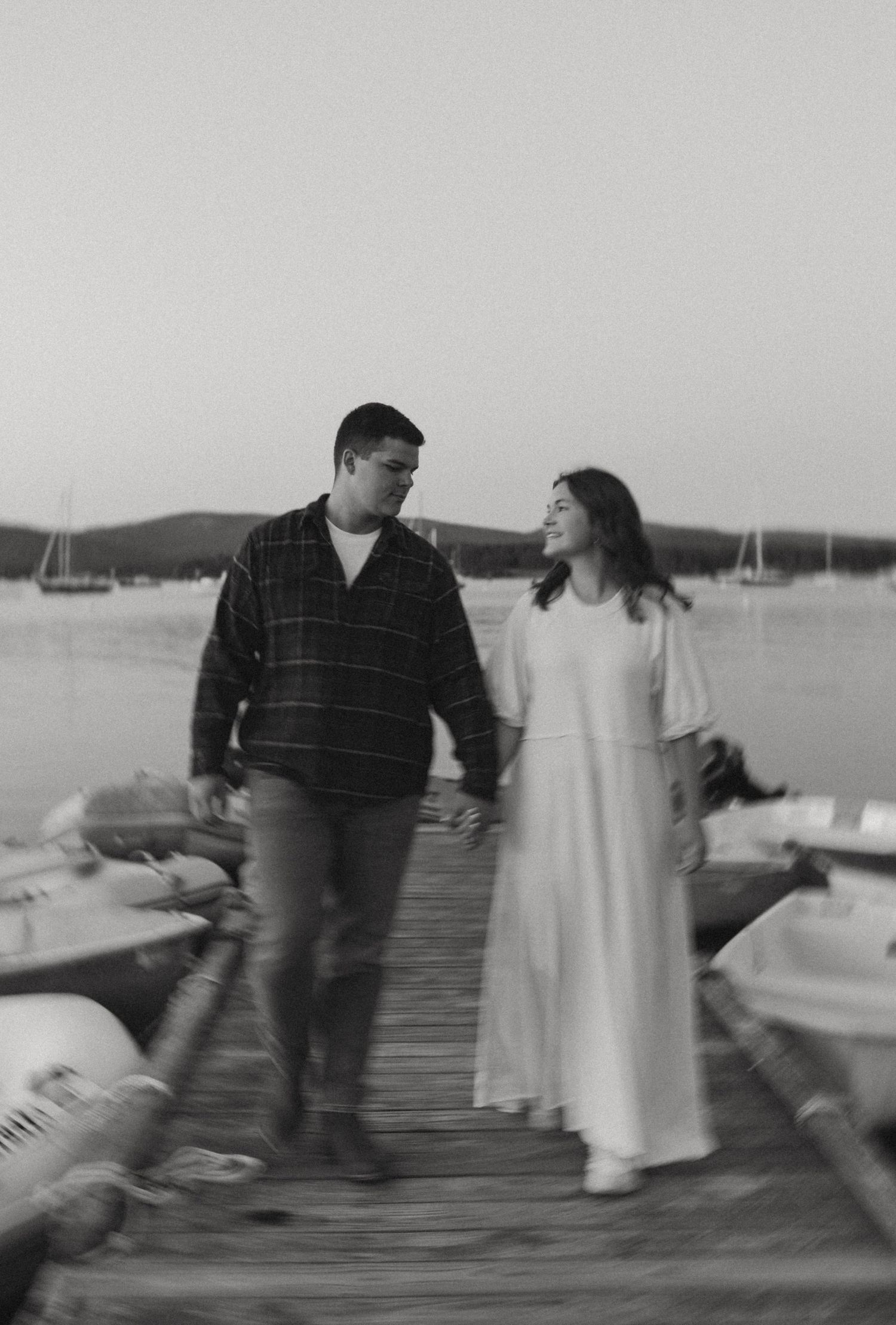 Black and white photo of couple walking hand in hand along a marina dock at sunset.