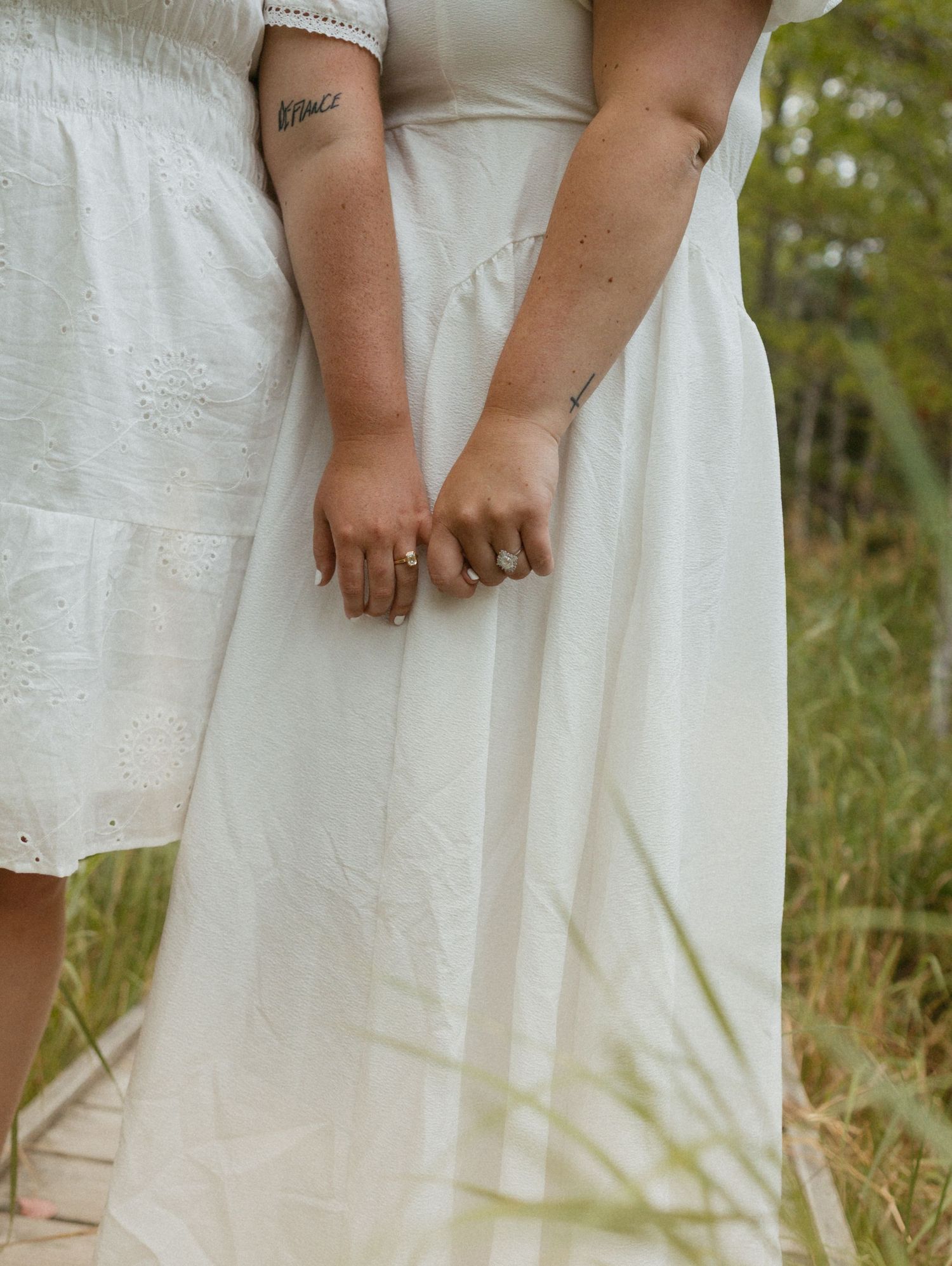 Two brides in white dresses hold hands outdoors, showing wedding rings and a delicate arm tattoo.