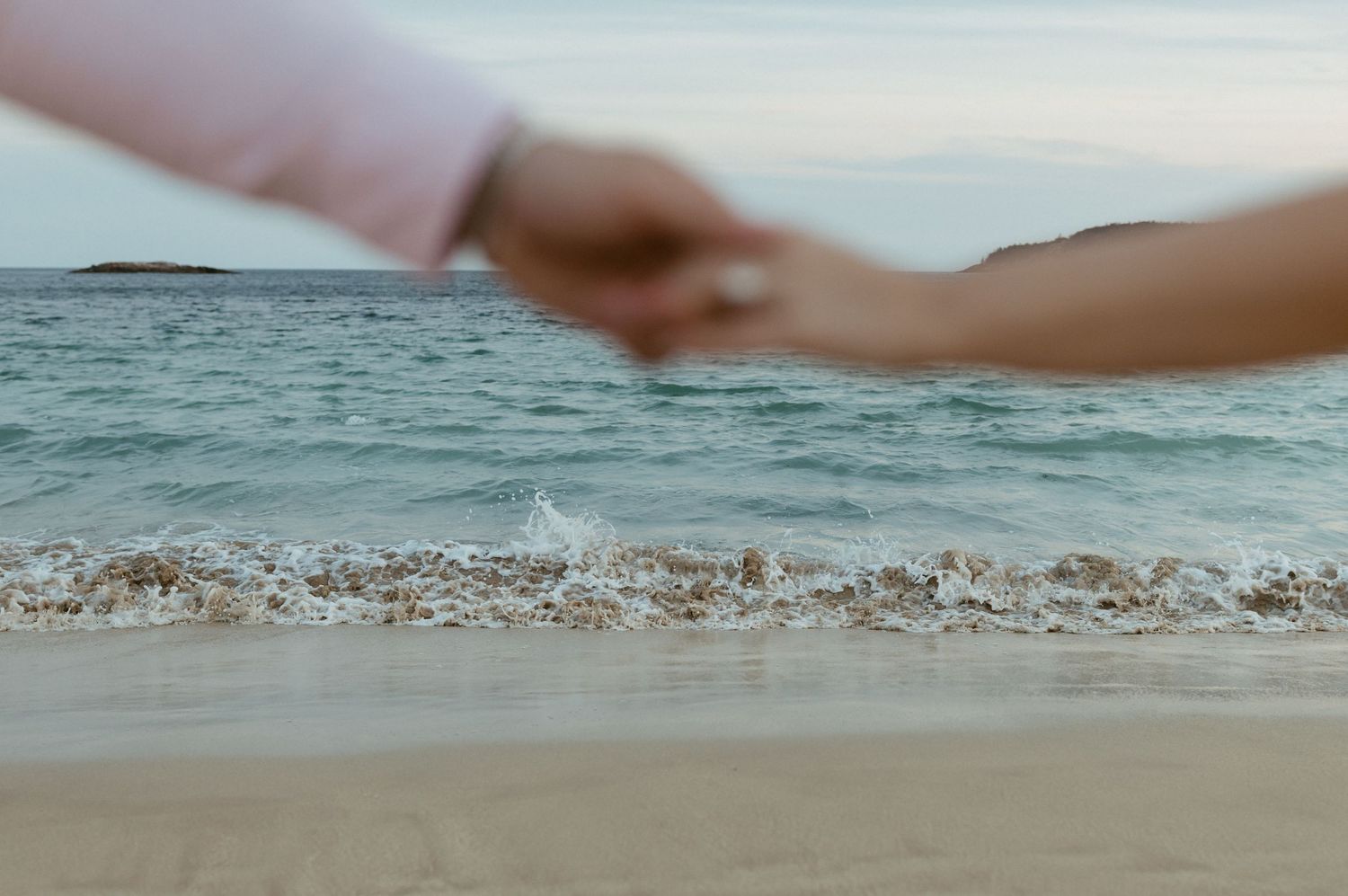Close-up of couple holding hands on sandy beach with ocean waves in background.