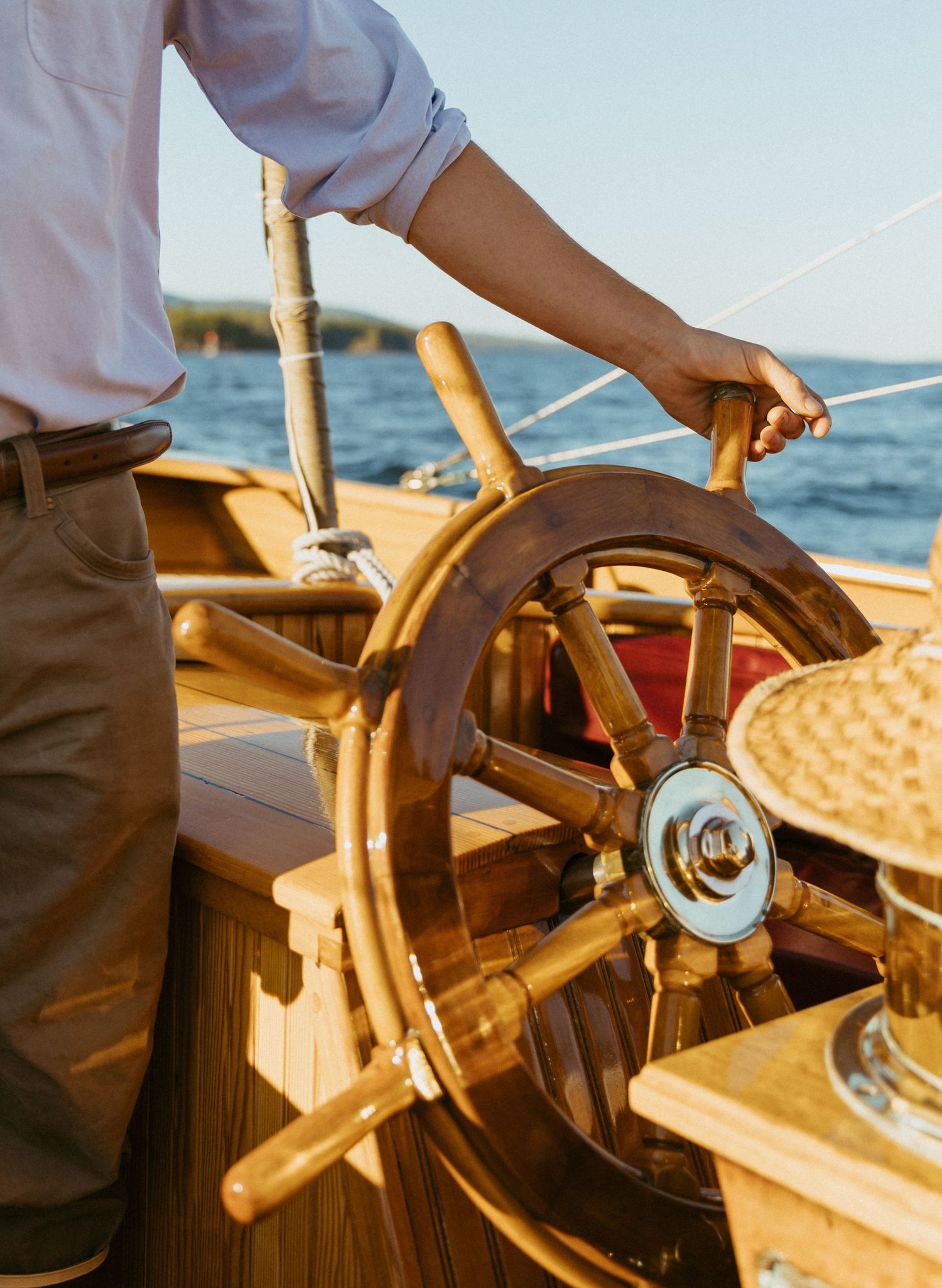 Person steers a classic wooden sailboat with a vintage wooden ship wheel on a sunny day at sea.
