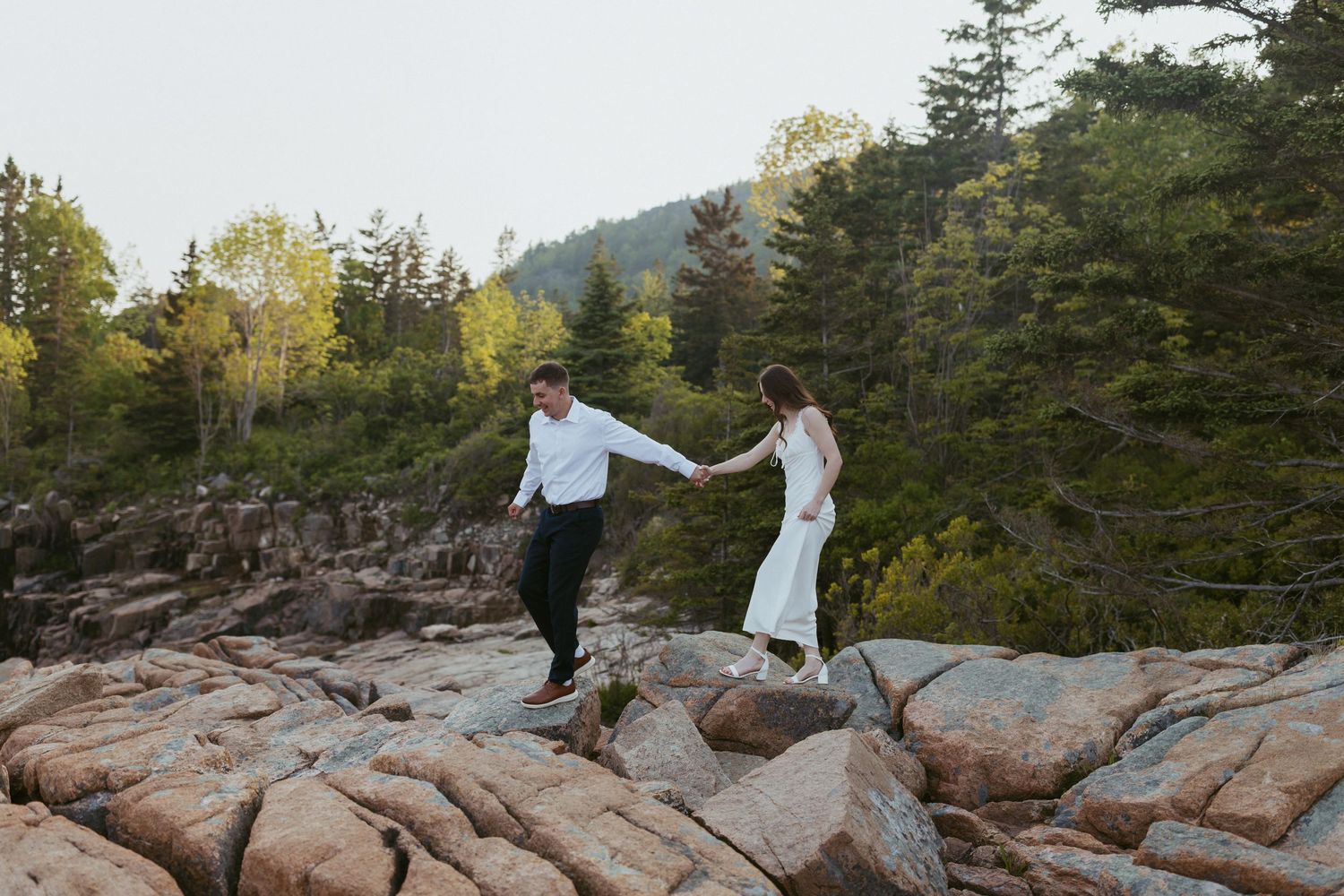 Bride and groom holding hands while walking on rocky coastal terrain surrounded by pine trees.