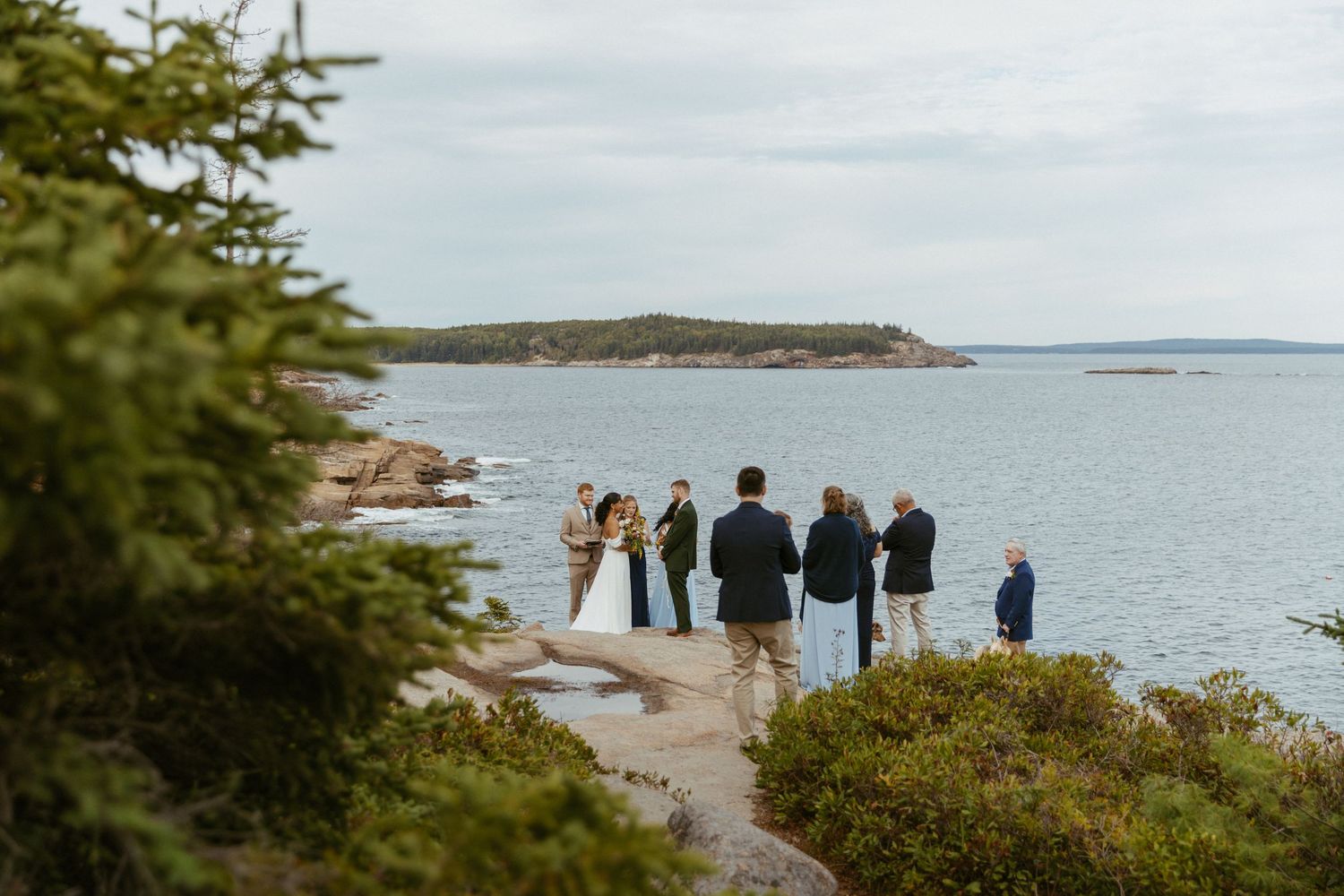 Small intimate outdoor wedding ceremony on coastal rocks overlooking a calm ocean bay.