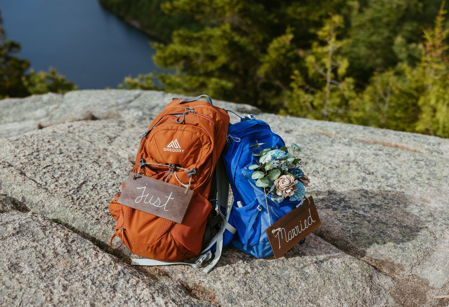 Two hiking backpacks labeled Just Married sit on a granite rock overlooking a forest lake.