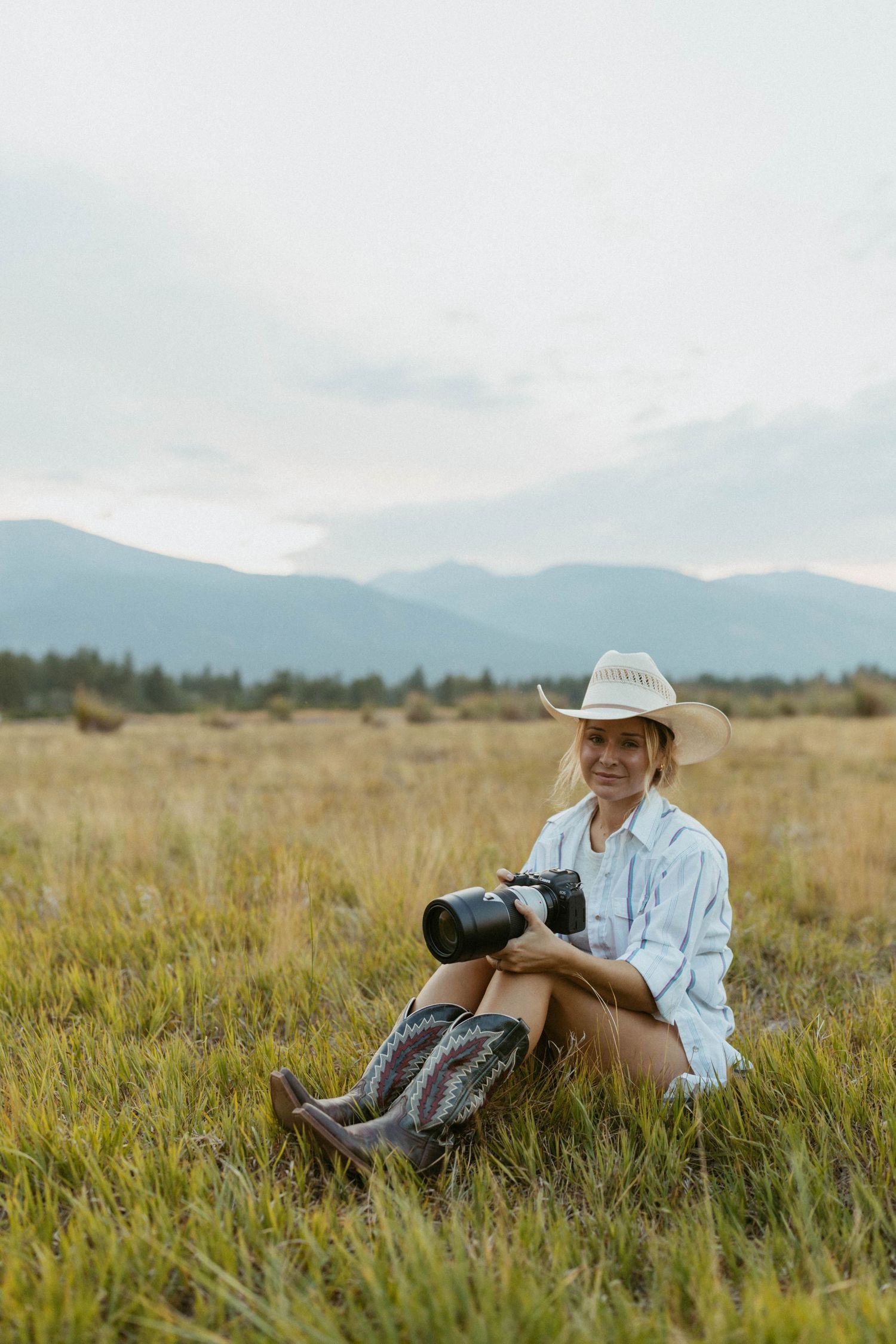Woman in cowboy hat and boots sits in grassy field holding camera with mountain backdrop.