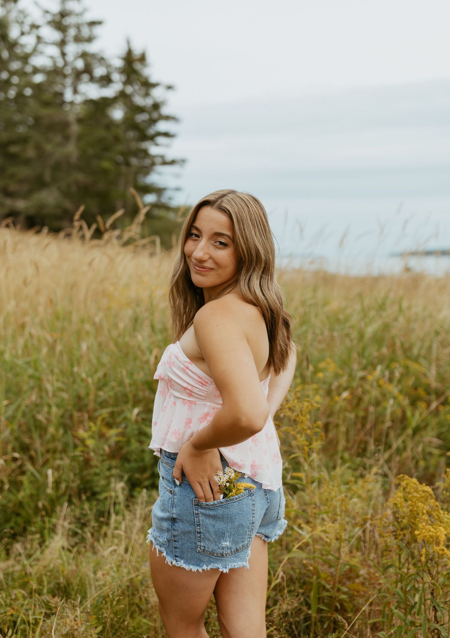 Young woman in pink top and denim shorts holding wildflowers in a golden meadow with trees and sky backdrop.