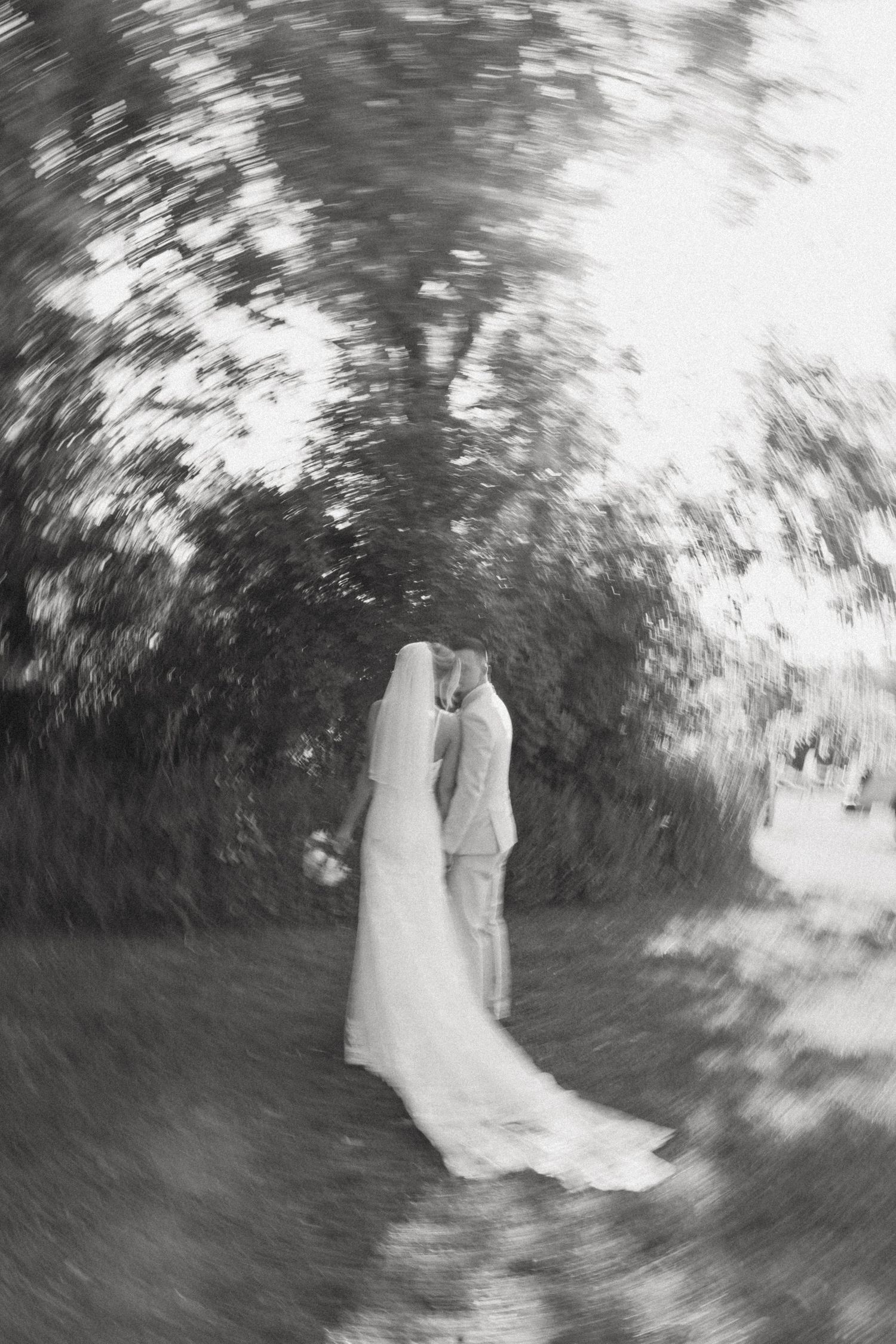 Black and white blurred photo of a bride and groom kissing under lush trees on their wedding day.