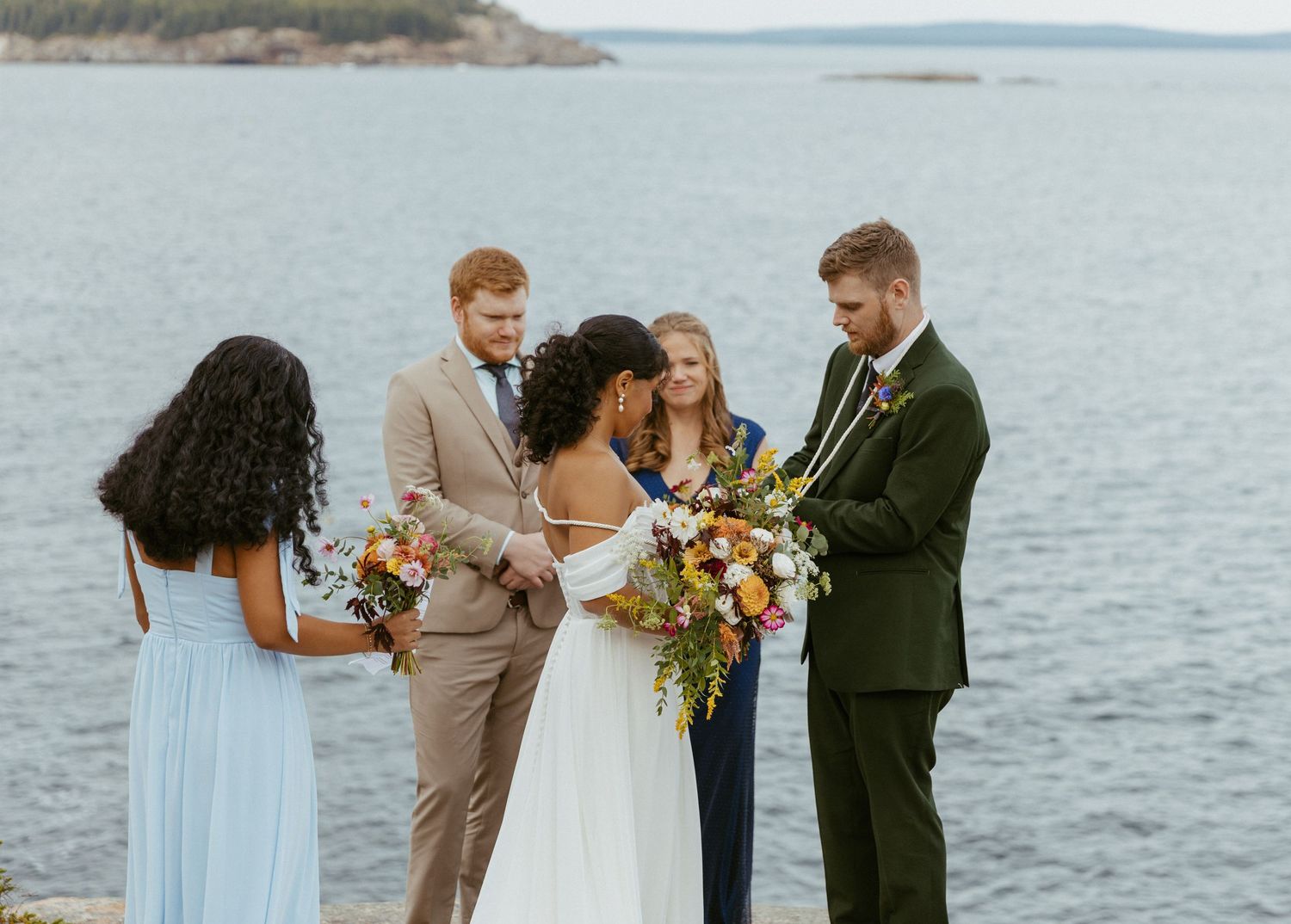 Bride and groom exchange vows with wedding party beside calm ocean waters.