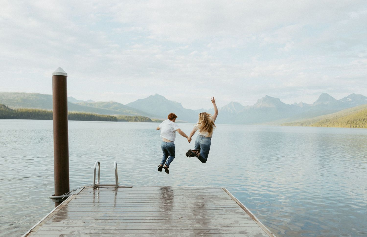 Couple joyfully jumping off a wooden dock into a serene mountain lake under a cloudy sky.