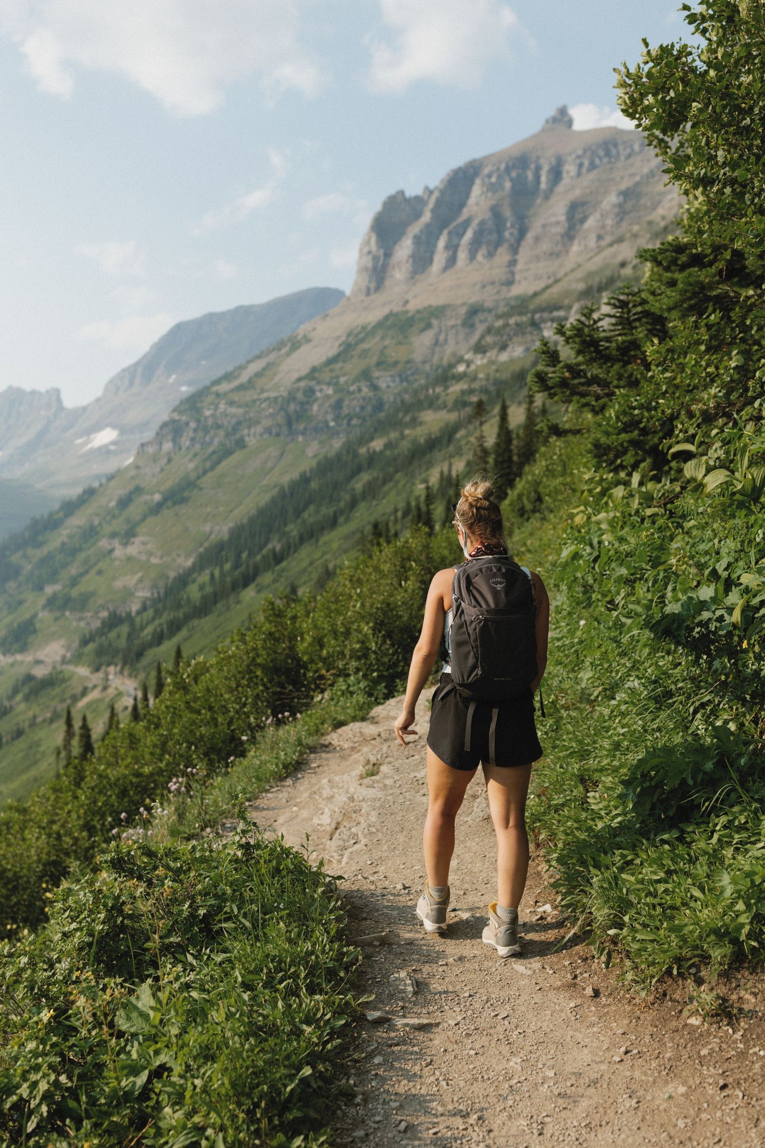 Female hiker with backpack walking mountain trail surrounded by lush greenery in Glacier National Park.