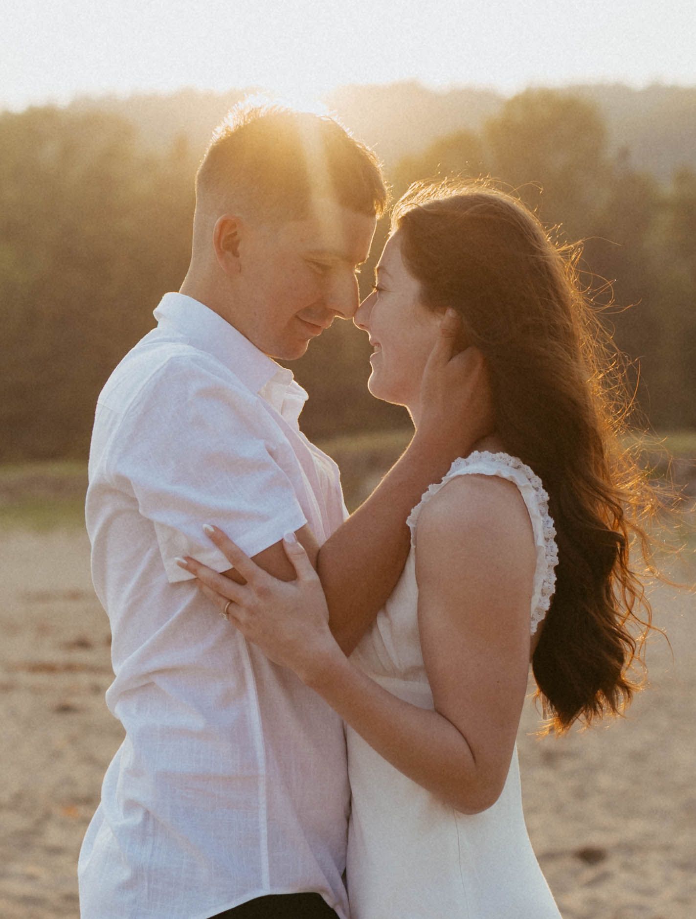 Couple nearly kissing in golden sunset light on Acadia sandy beach, embracing romantically.