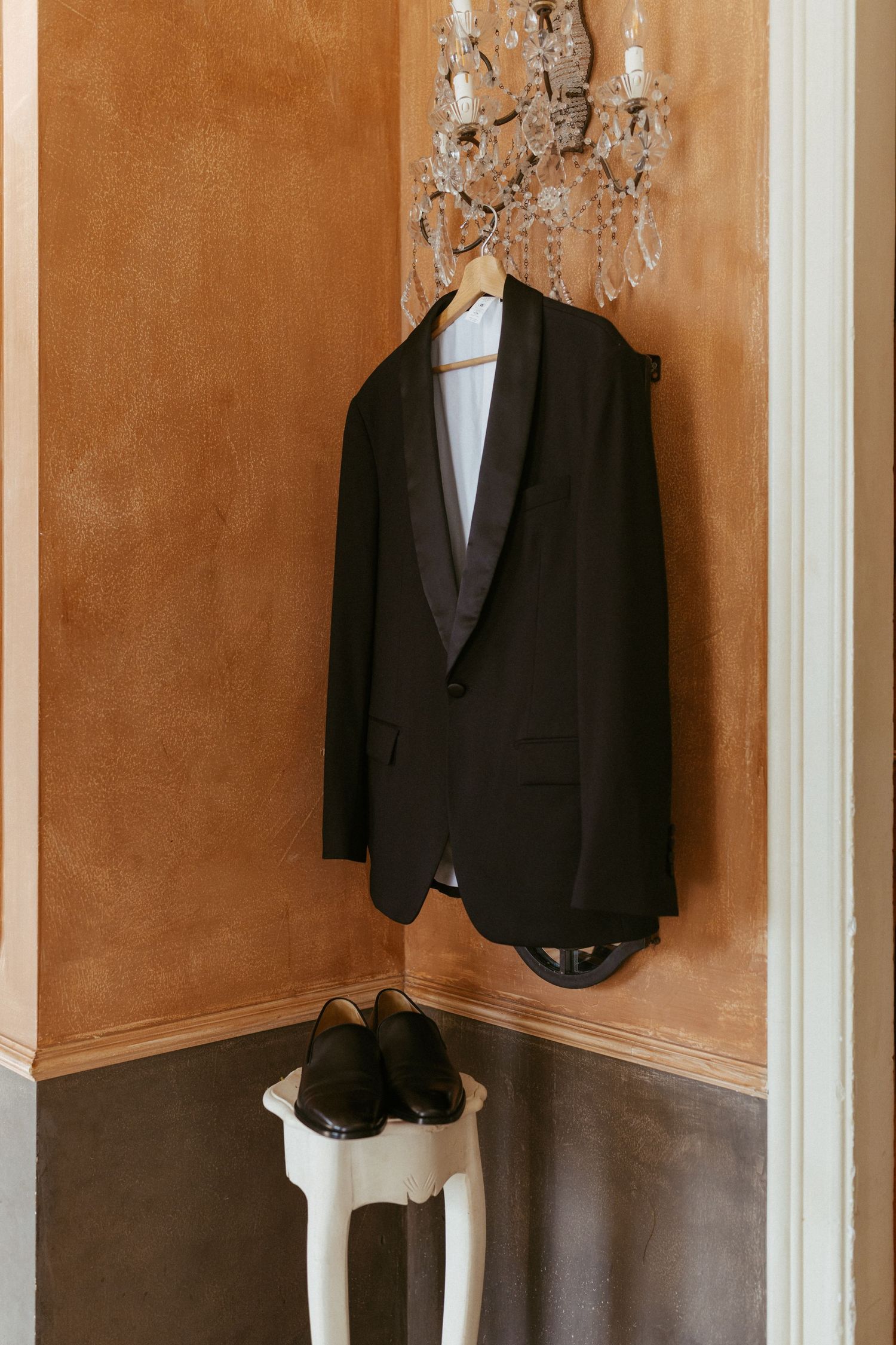 Black tuxedo jacket hanging on crystal chandelier with dress shoes on white stool below.