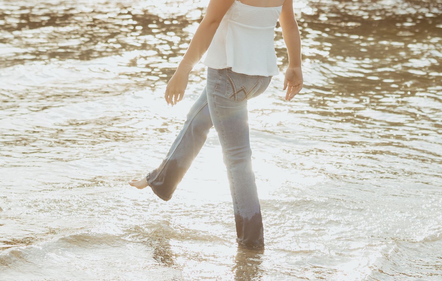 Person in white top and light jeans wading through shallow ocean waves on a sandy beach.