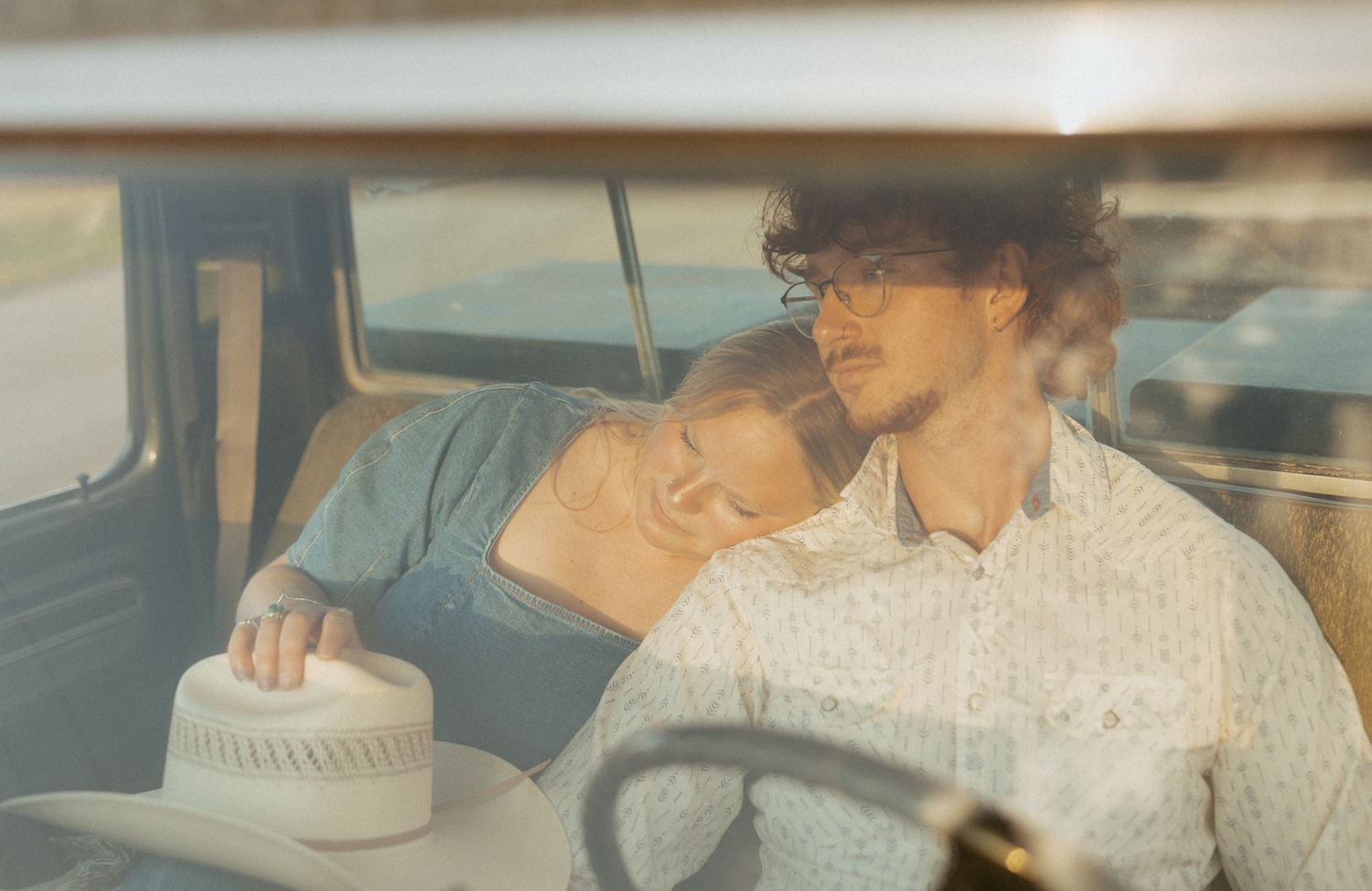Romantic couple resting together inside vintage truck cab, woman leaning on mans shoulder holding a cowboy hat.