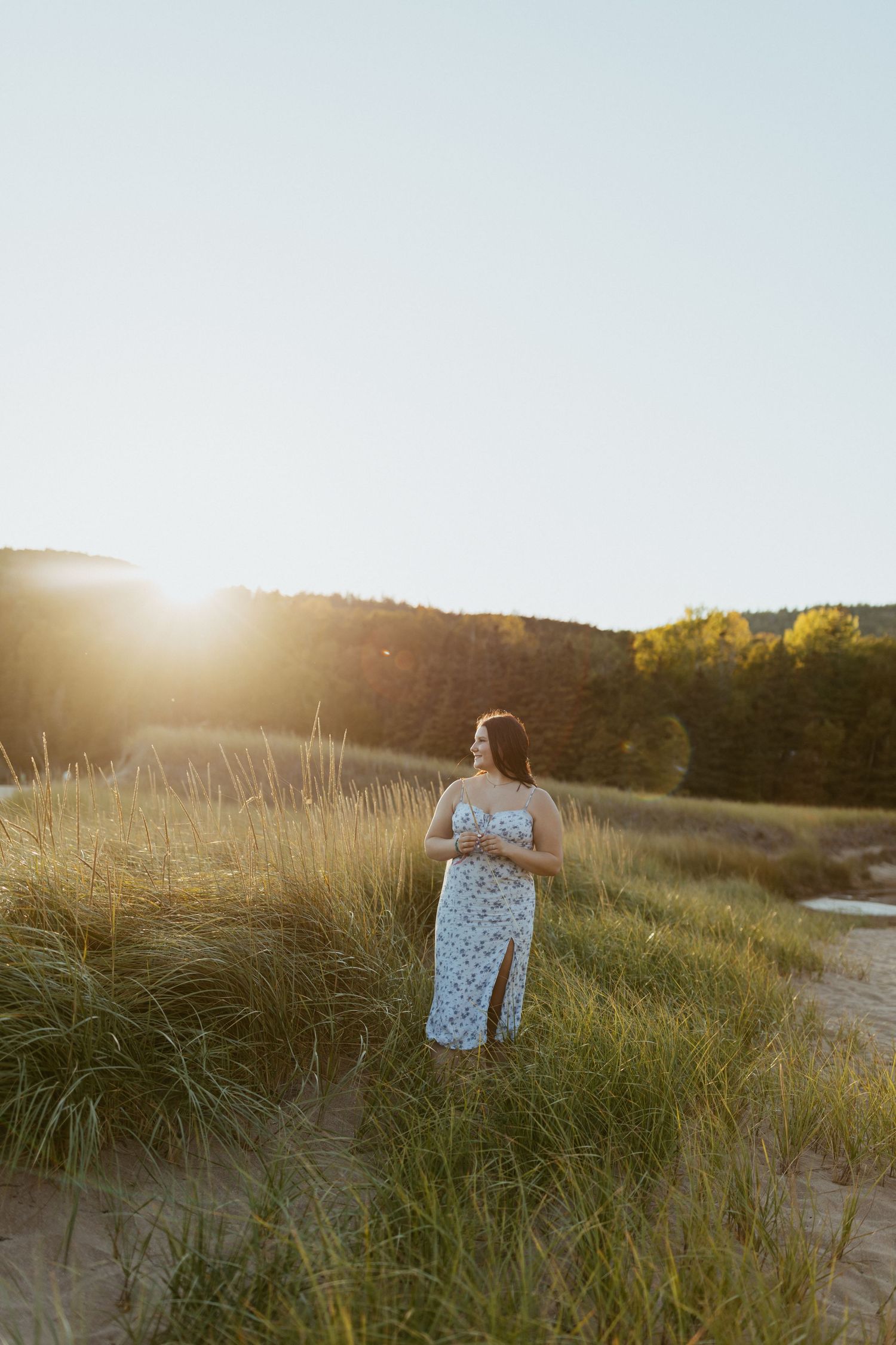 Woman in floral dress standing among sand dunes at golden hour with sunlight glowing behind green hills.