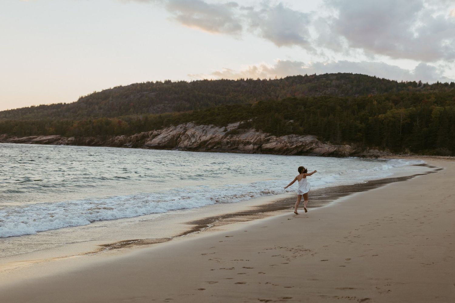 A child plays along a sandy beach shore at sunset with forested hills in the background.