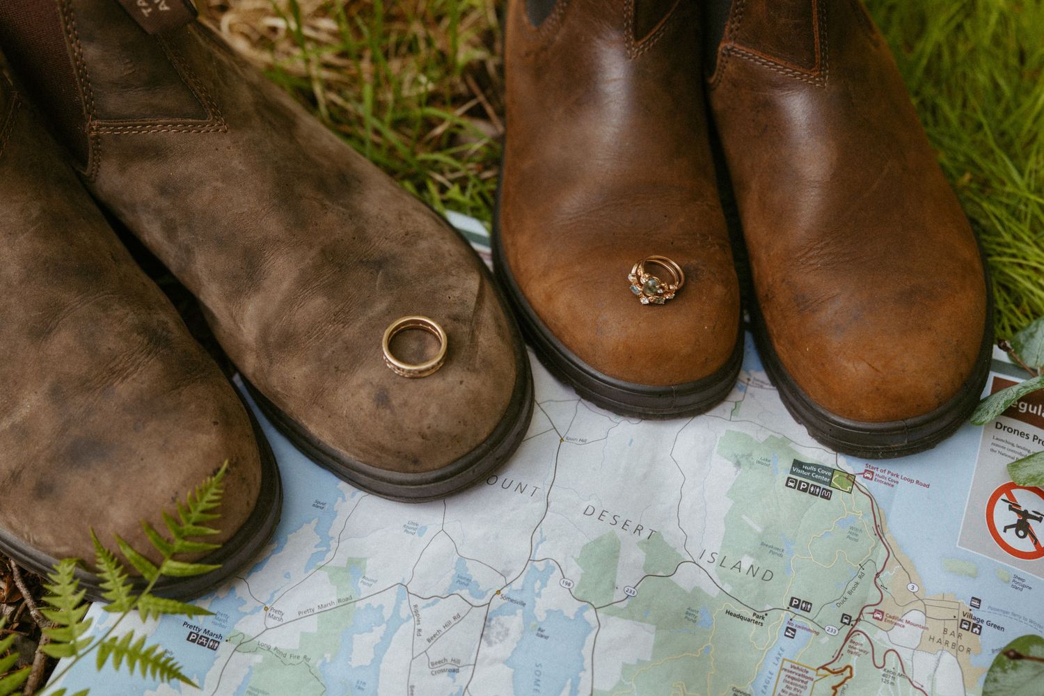 Two brown boots with wedding rings resting on top of a map, surrounded by green grass and ferns.
