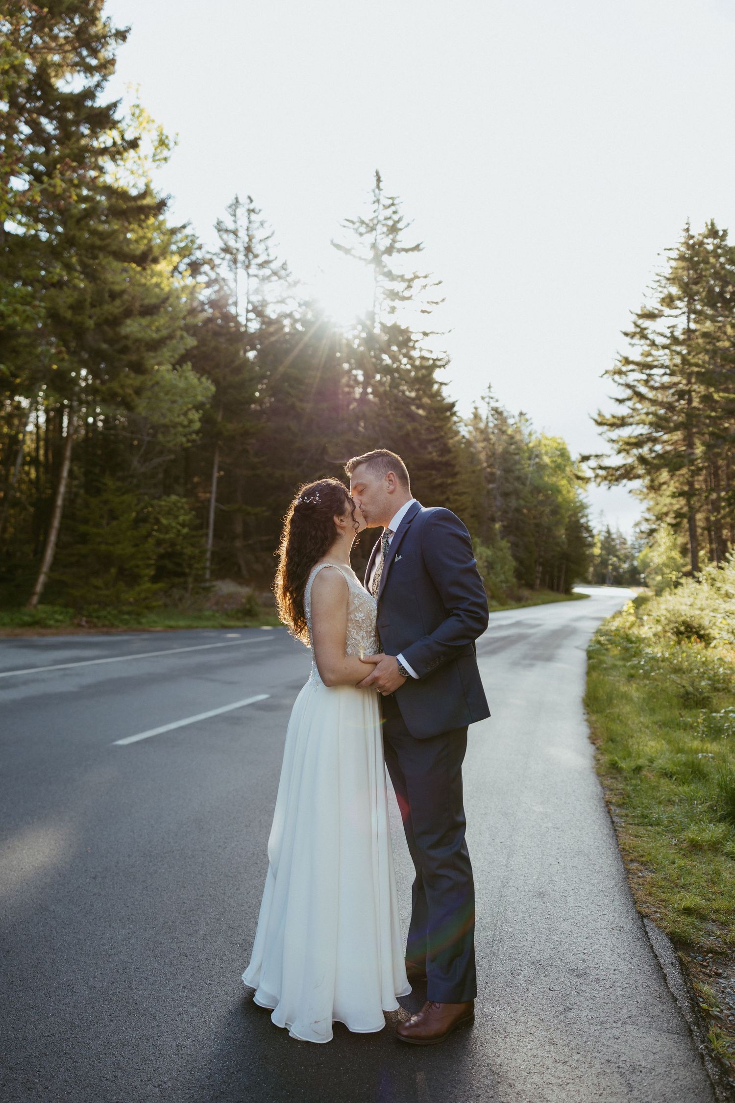 Bride and groom kissing on a sunlit forest road, wearing wedding attire, surrounded by tall pine trees.