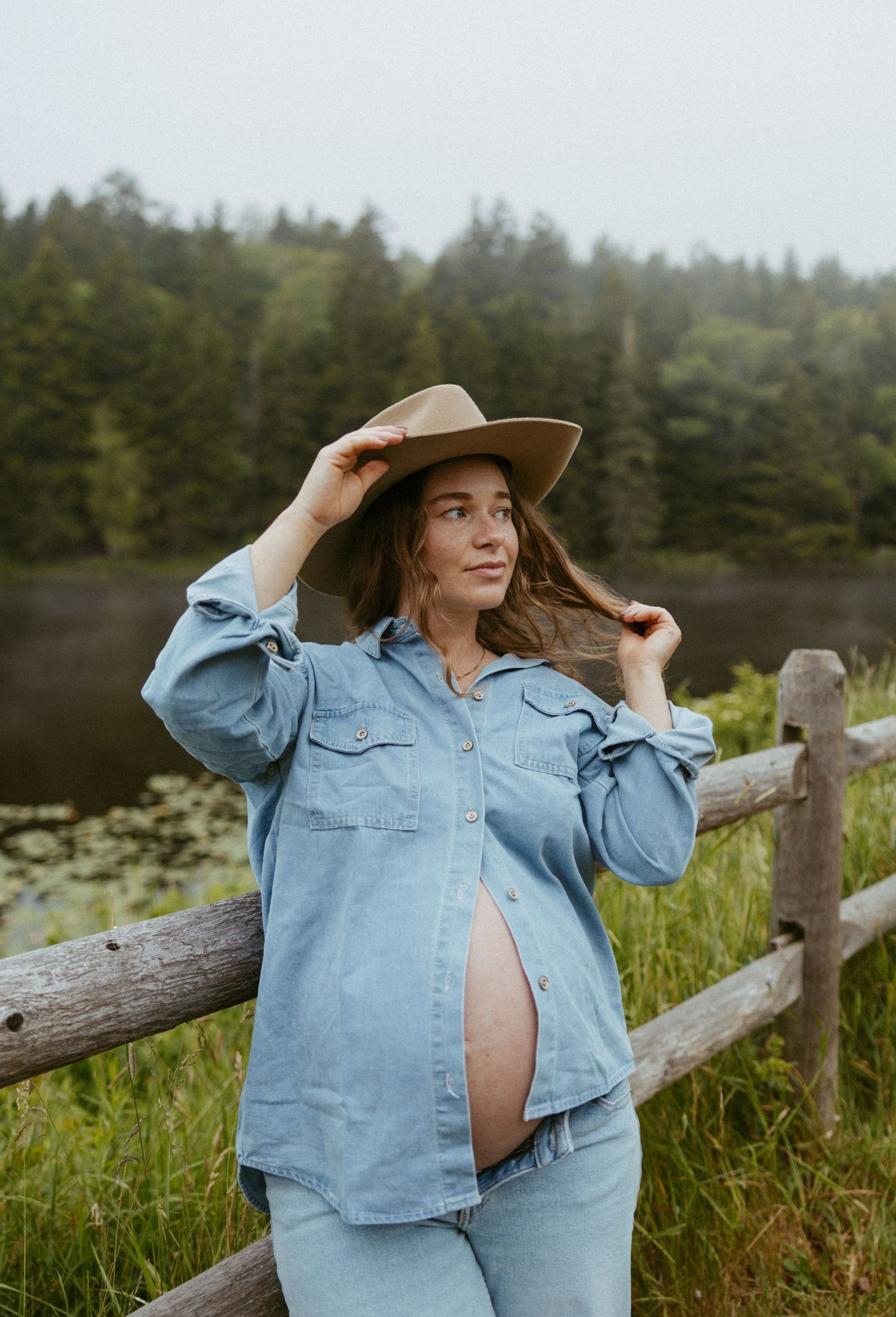 Pregnant woman in denim outfit and tan cowboy hat poses by rustic fence near lake and forest.
