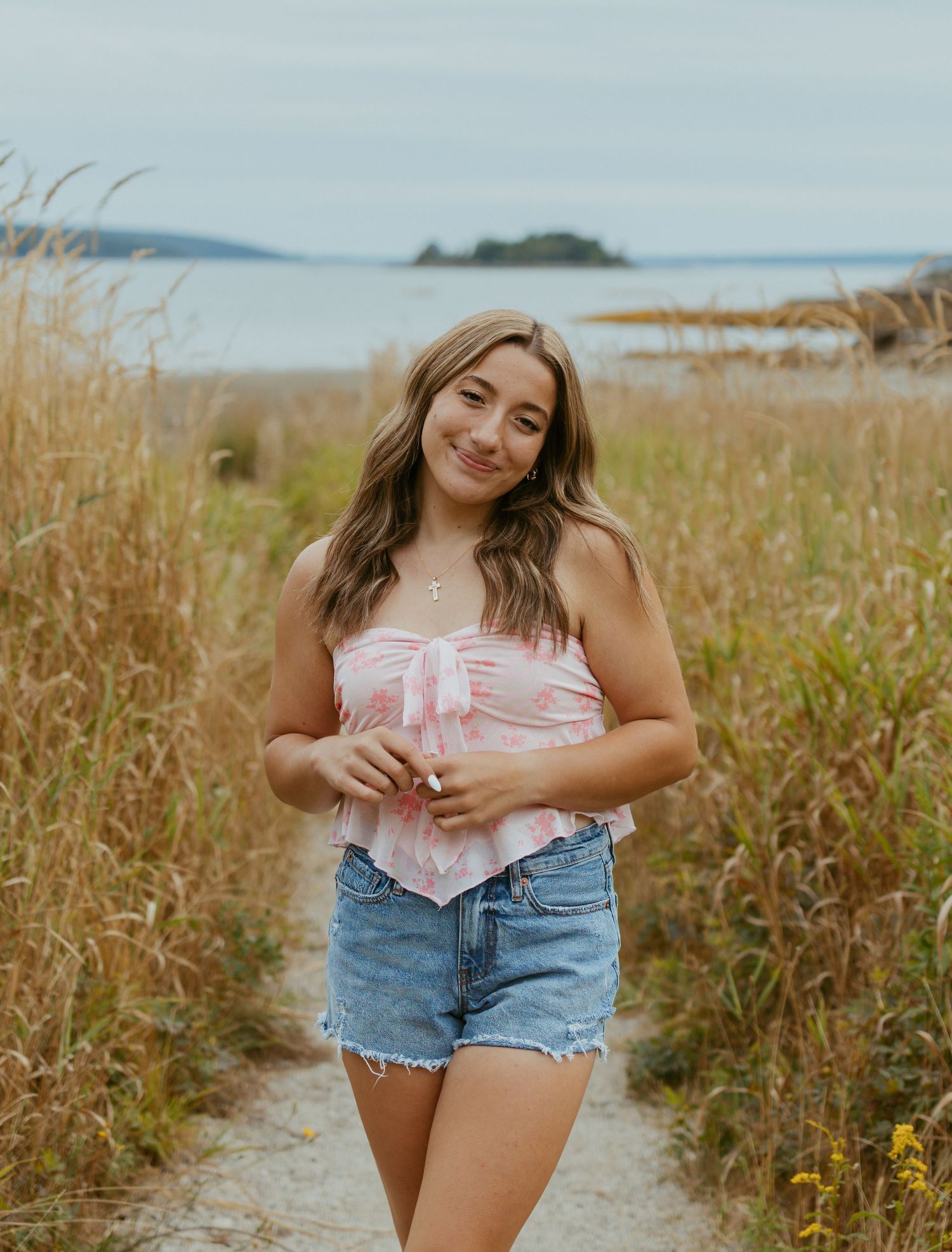 Young woman in pink top and denim shorts smiling on a coastal trail surrounded by golden tall grass.