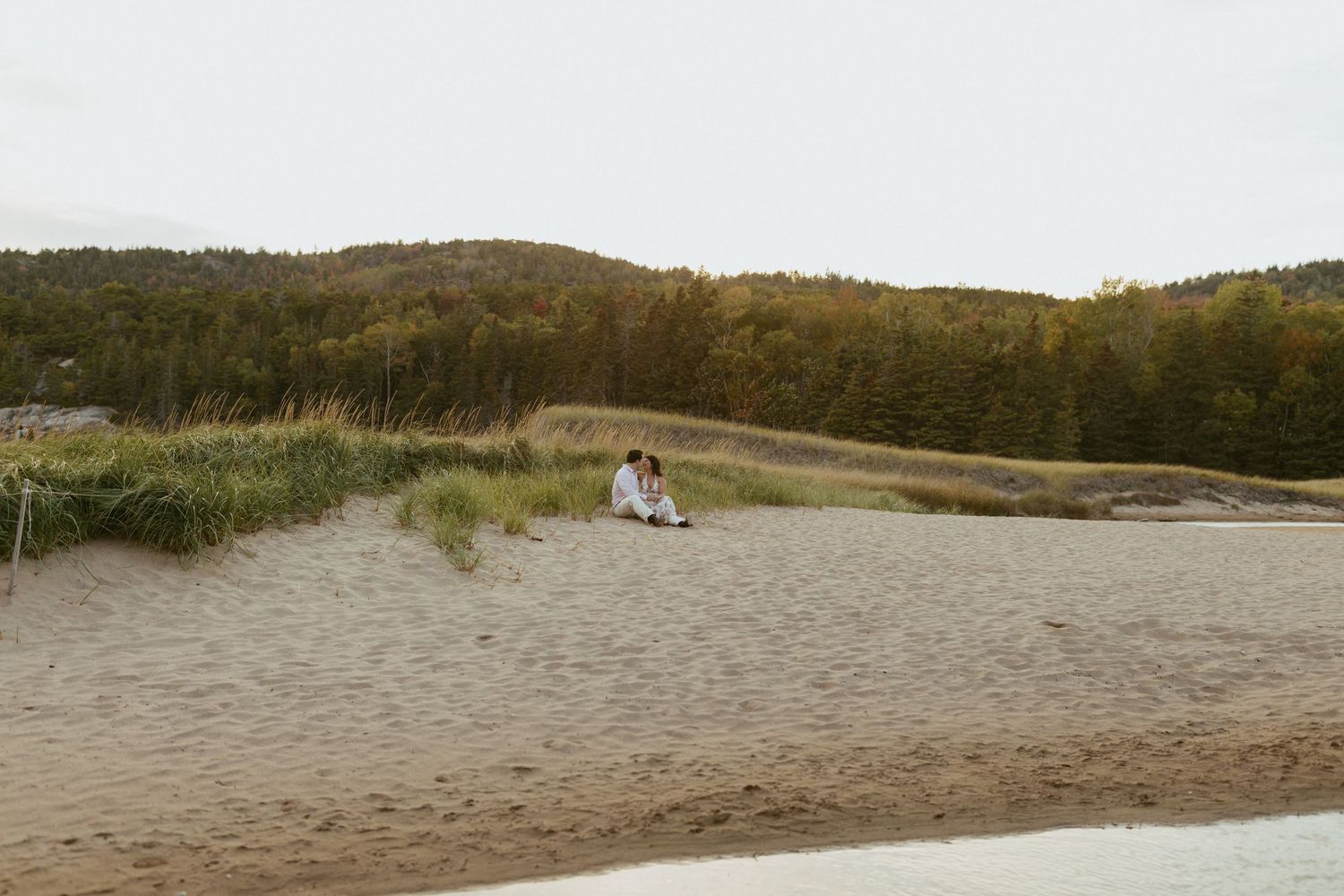 Couple sitting close together on a wide sandy beach surrounded by dunes and forest hills.